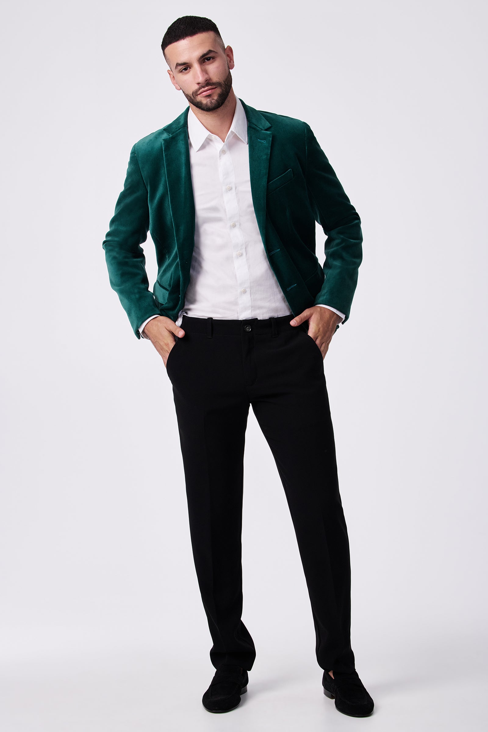 A man with short hair and a trimmed beard stands against a plain background, wearing black Alex Slim Trousers in drapey suiting, a green velvet blazer, white dress shirt, and black shoes. One hand is in his pocket and his posture is relaxed.