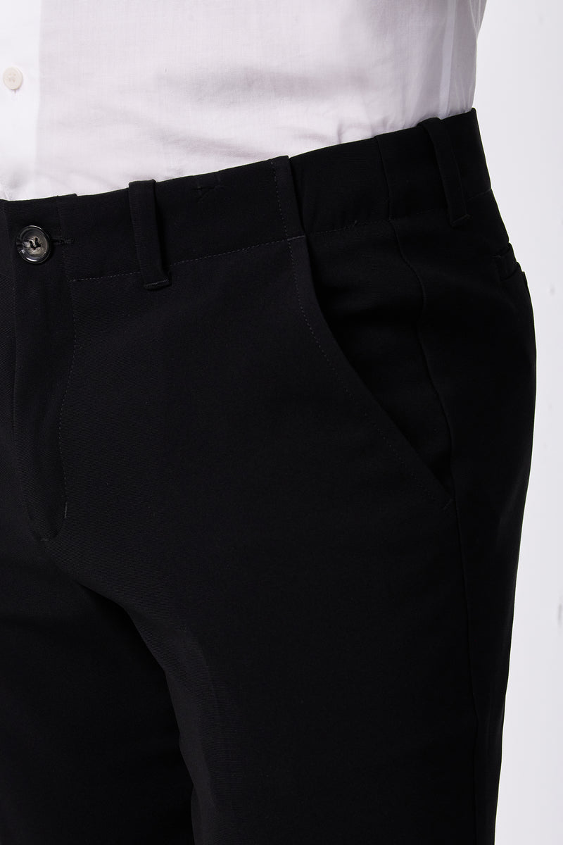 A person wearing the Alex Slim Trouser - Drapey Suiting in black with a white shirt, shown from waist to mid-thigh, one hand in the trouser pocket.