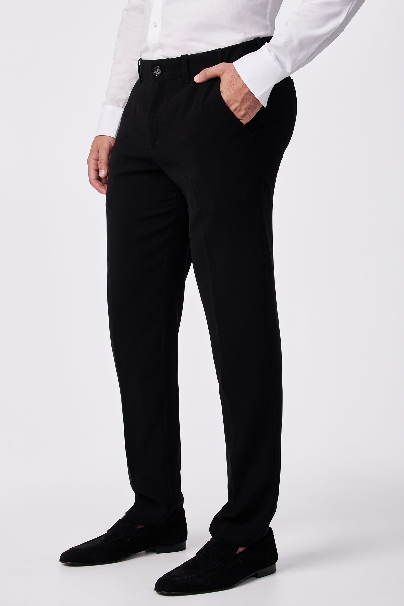 A man wearing the Alex Slim Trouser - Drapey Suiting, paired with a white button-up shirt and black dress shoes, stands with one hand in his pocket against a plain white background.