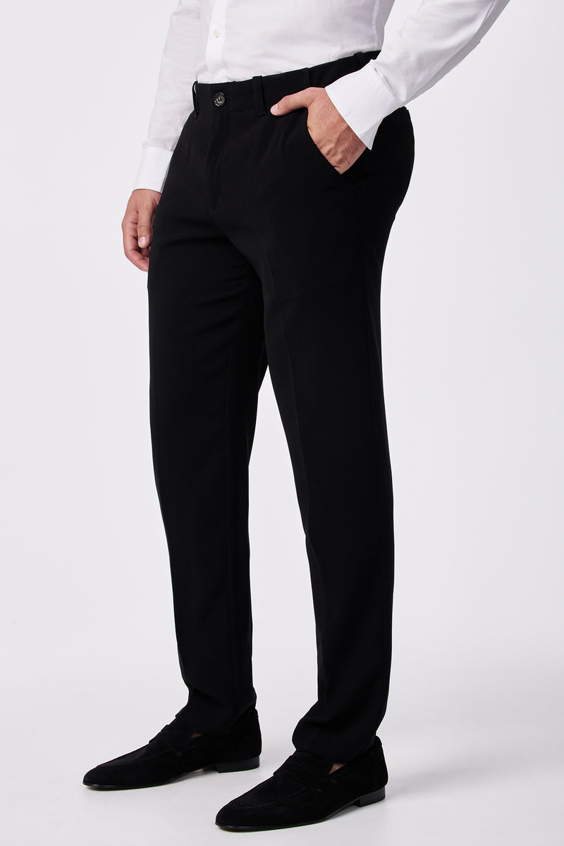 A man wearing the Alex Slim Trouser - Drapey Suiting, paired with a white button-up shirt and black dress shoes, stands with one hand in his pocket against a plain white background.