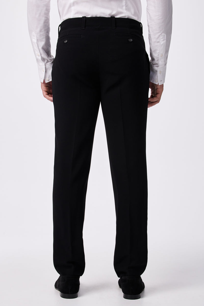 A person wearing a white dress shirt and the Alex Slim Trouser - Drapey Suiting in black is standing with their back to the camera, highlighting the back view of the trousers.