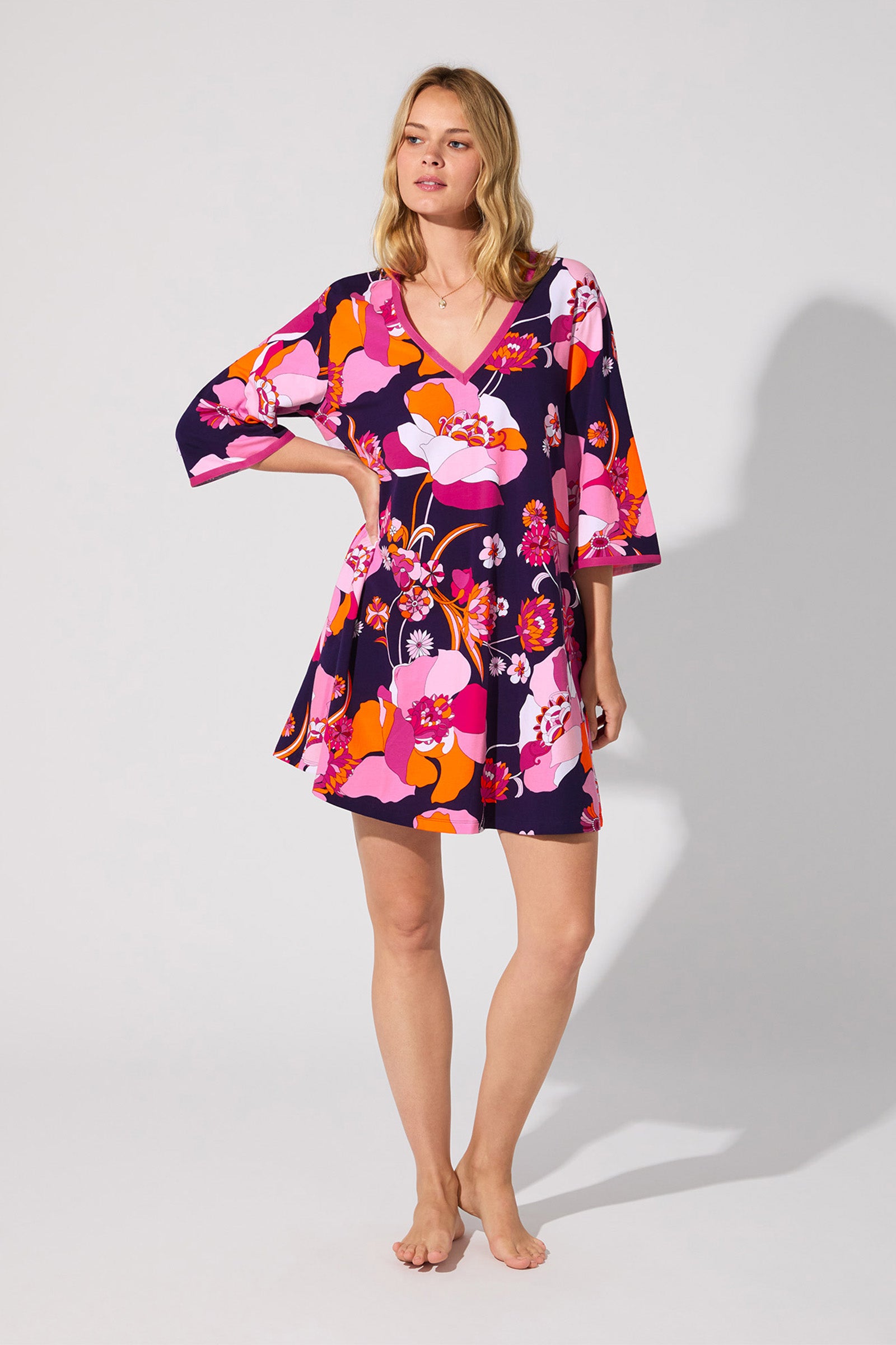 A woman stands barefoot against a light background in the Floral Medley Caftan, a loose dress with bold pink, orange, white, and purple floral patterns and wide sleeves. She poses with one hand on her hip, gazing slightly to the side.