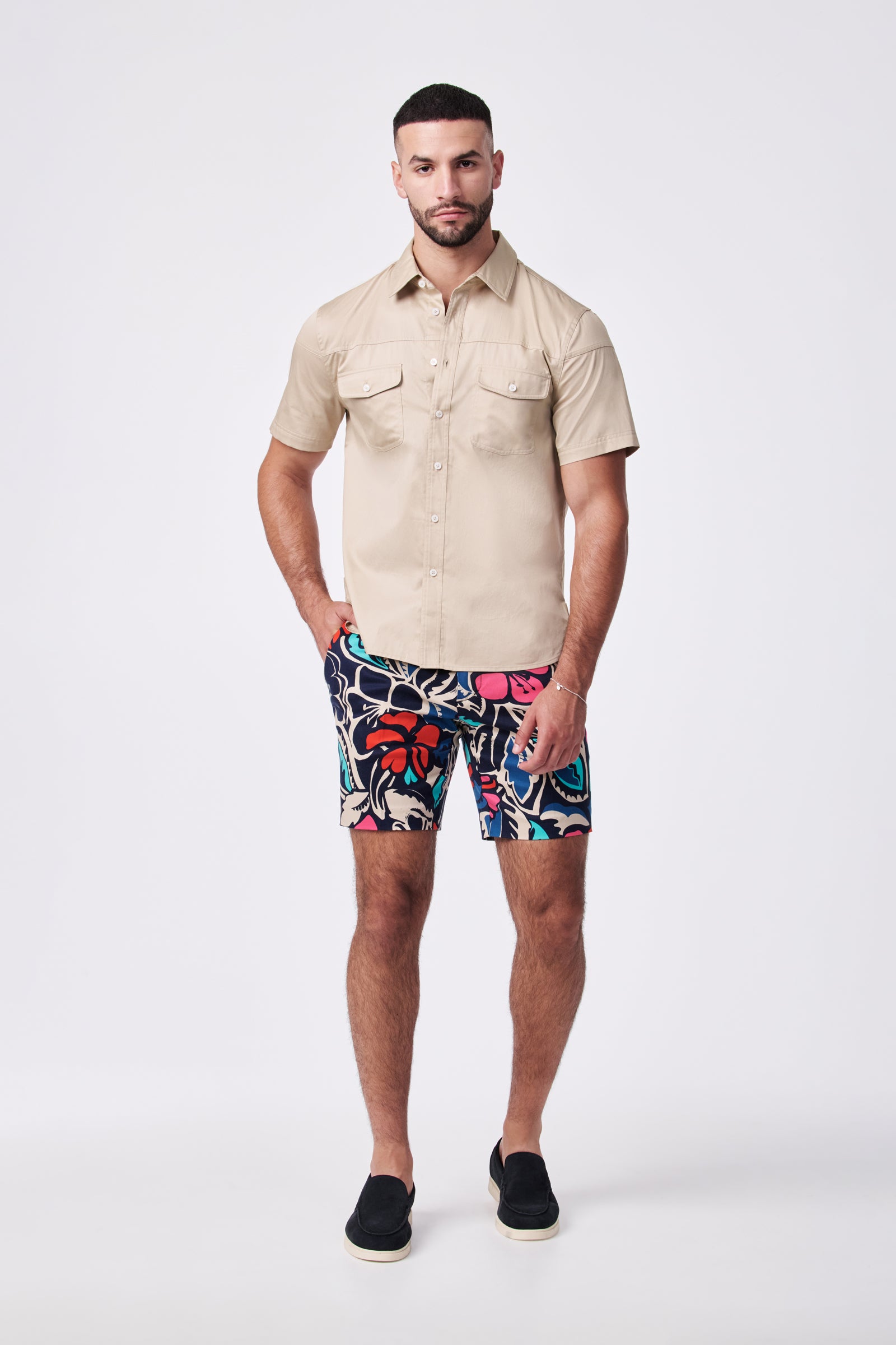 A man stands against a white background wearing the Lawrence Short, a beige short-sleeve button-up shirt, and black slip-on shoes. His hands are in his pockets, and he faces forward.