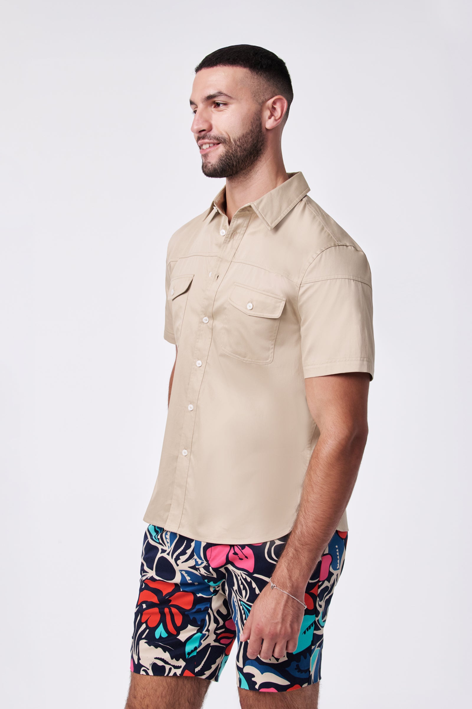A bearded man is wearing the Shelton Ss Shirt, a beige short-sleeve button-up, paired with colorful floral shorts. He's standing, smiling, and facing slightly to the side against a plain white background.
