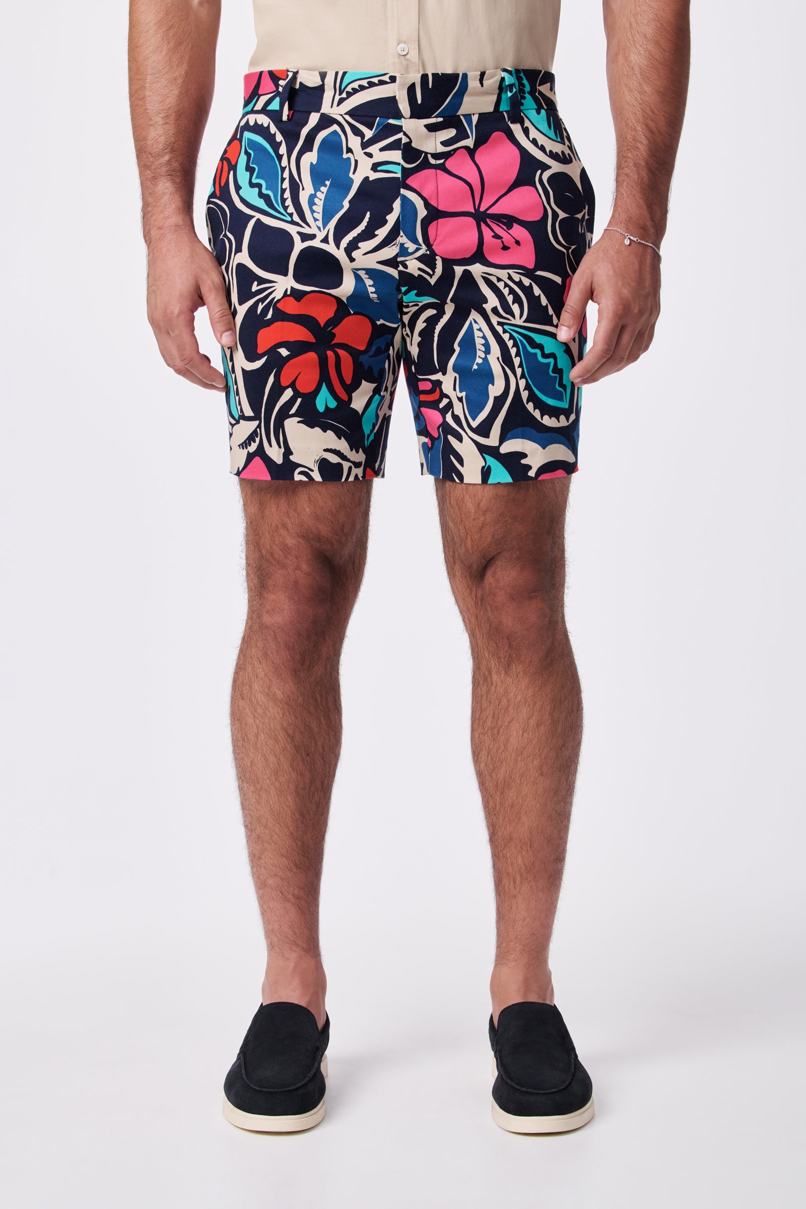 A man models the Lawrence Short in a colorful floral pattern, paired with a beige tucked-in shirt and black slip-on shoes. The image captures his legs and torso against a plain white background.