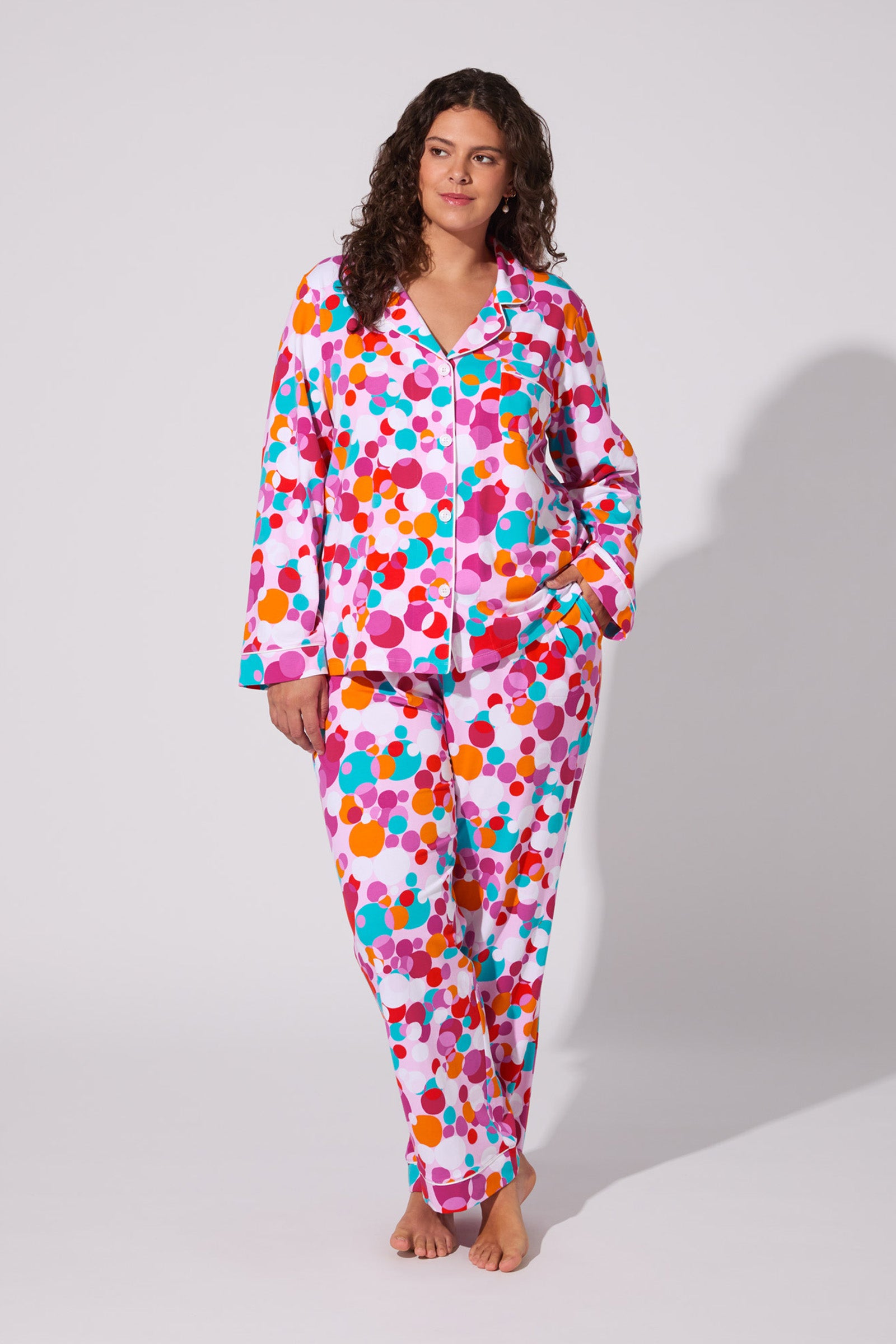 A woman with wavy brown hair stands barefoot against a light background, wearing the Bubble Dots Classic Pj Set—a colorful, long-sleeved pajama set featuring a bright multicolored polka-dot pattern. She looks to the side with one hand in her pocket.