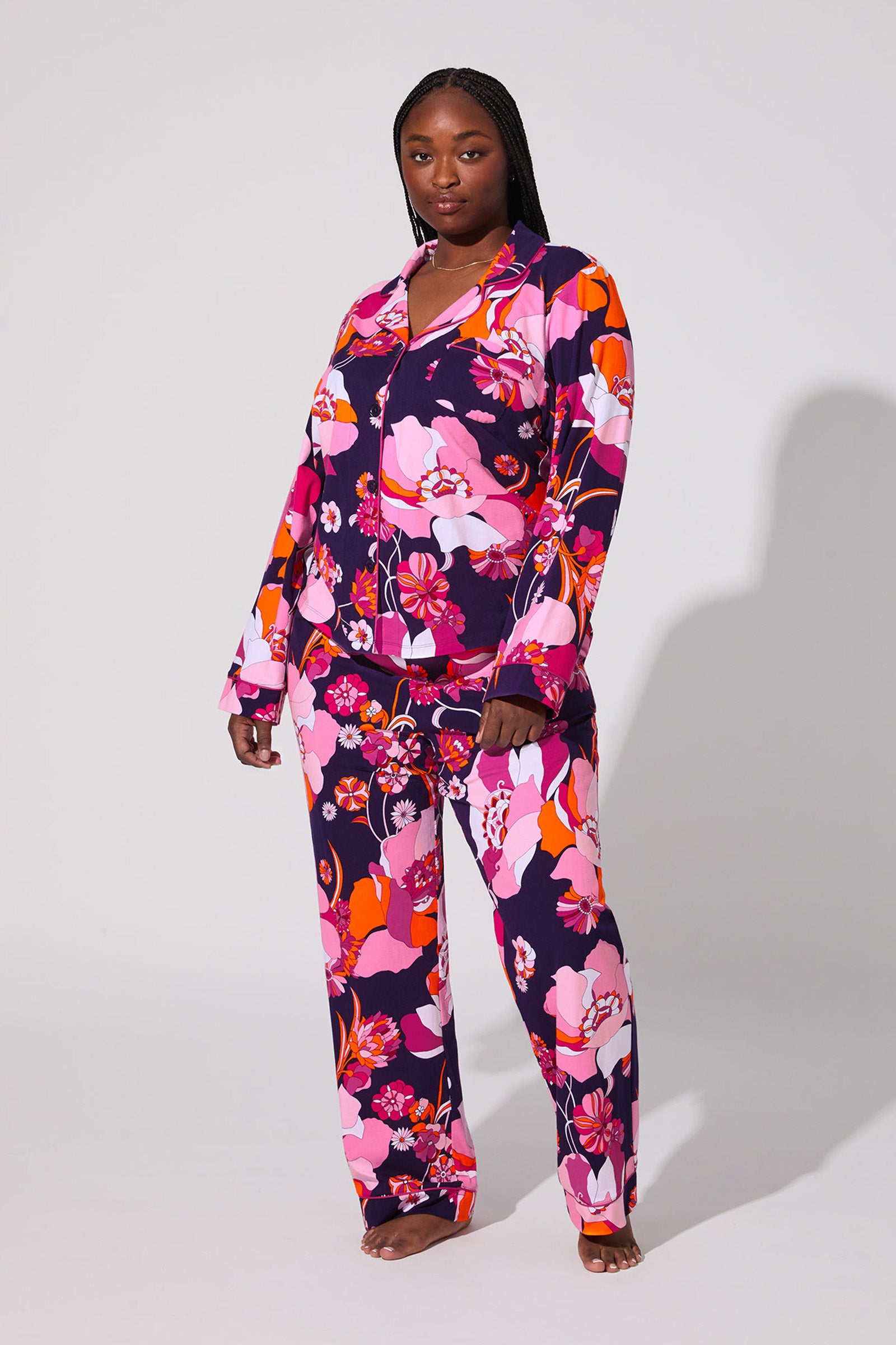 A woman poses confidently barefoot in the Floral Medley Classic Pj Set, featuring a bold floral pattern in shades of pink, orange, and purple on a dark background, against a plain light backdrop.