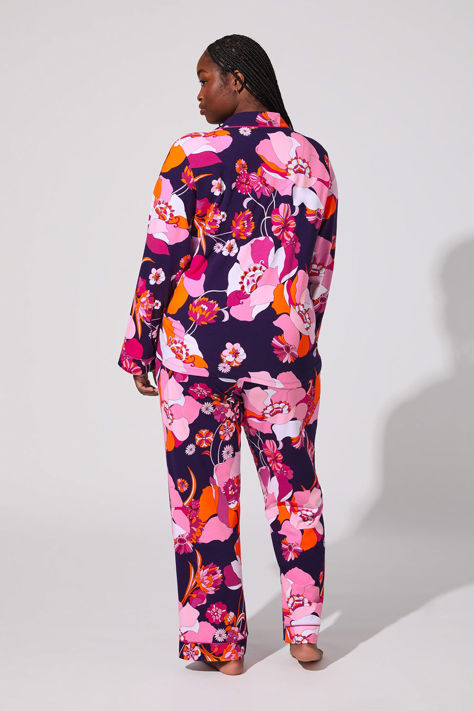 A person stands barefoot facing away, wearing the Floral Medley Classic Pj Set featuring a vibrant floral pattern in pink, orange, white, and purple against a plain light background.