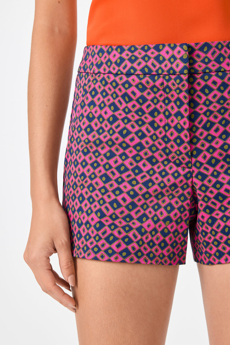 Someone wearing Corbin 2 Shorts - Himari Herringbone, featuring a vibrant pink, purple, and blue geometric diamond pattern with an orange top. Only the lower torso and one arm are visible.