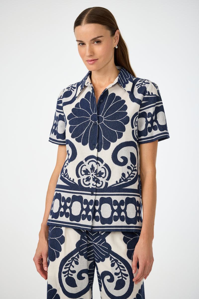 A woman with straight brown hair wears the Abba Top in Fuyuko Fil Coupe, featuring a bold navy and white floral and geometric pattern, against a plain light background.