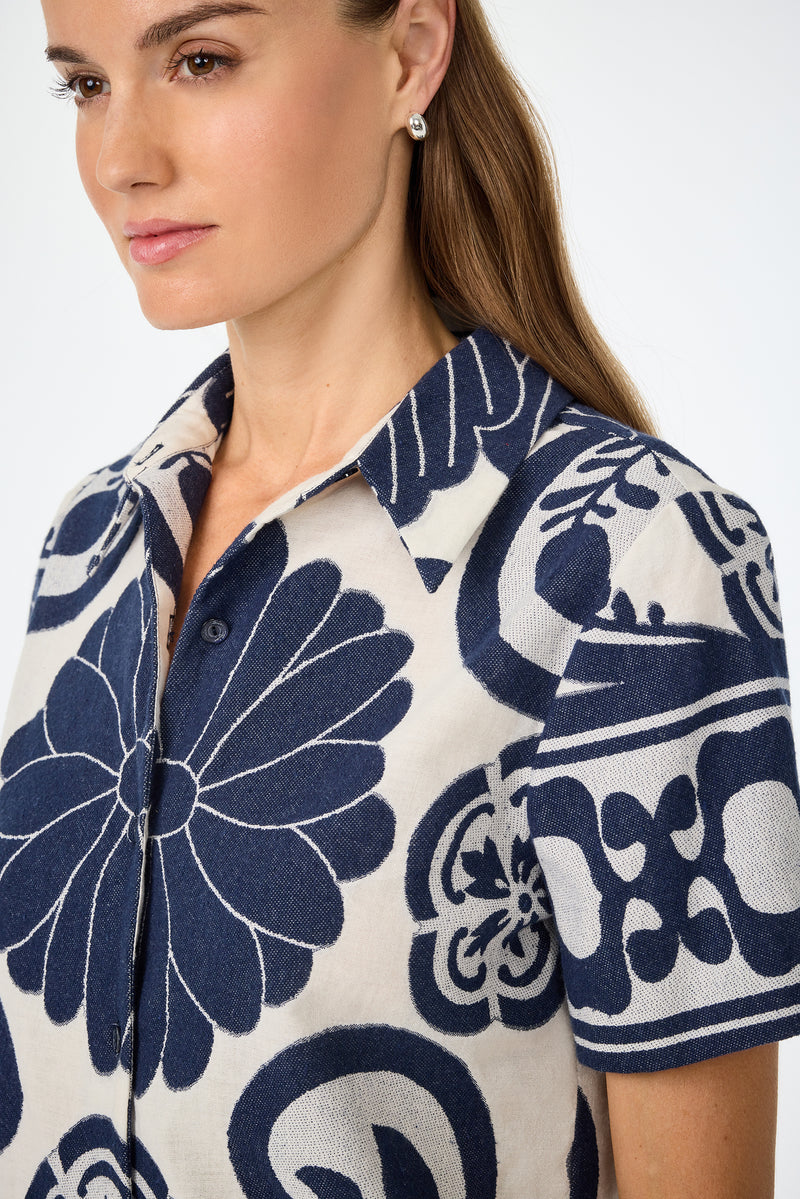 A woman with long brown hair wears the Abba Top - Fuyuko Fil Coupe, featuring a bold navy blue and white floral geometric pattern, as she looks slightly to the side against a plain light background.