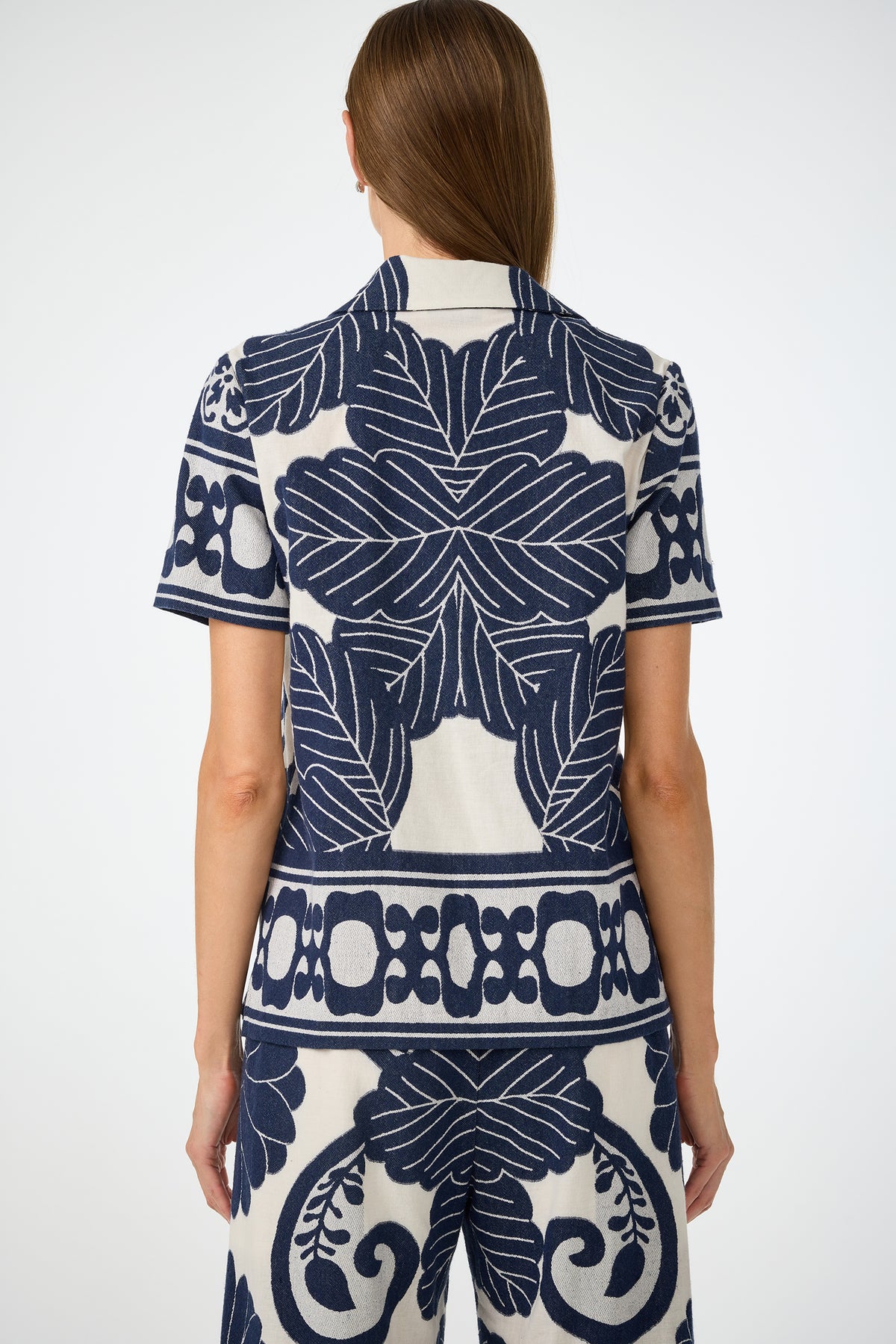 Someone with long brown hair, seen from behind, wears the Abba Top - Fuyuko Fil Coupe, a short-sleeve shirt with bold navy blue and white floral and geometric patterns, paired with matching pants.