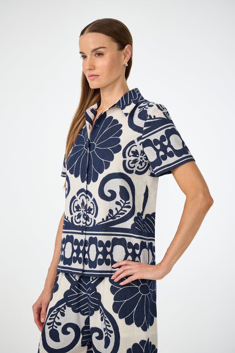 A woman with straight brown hair wears the Abba Top - Fuyuko Fil Coupe, a short-sleeved, collared shirt featuring a bold navy blue floral and geometric pattern on a white background. She stands sideways, looking ahead with a neutral expression.