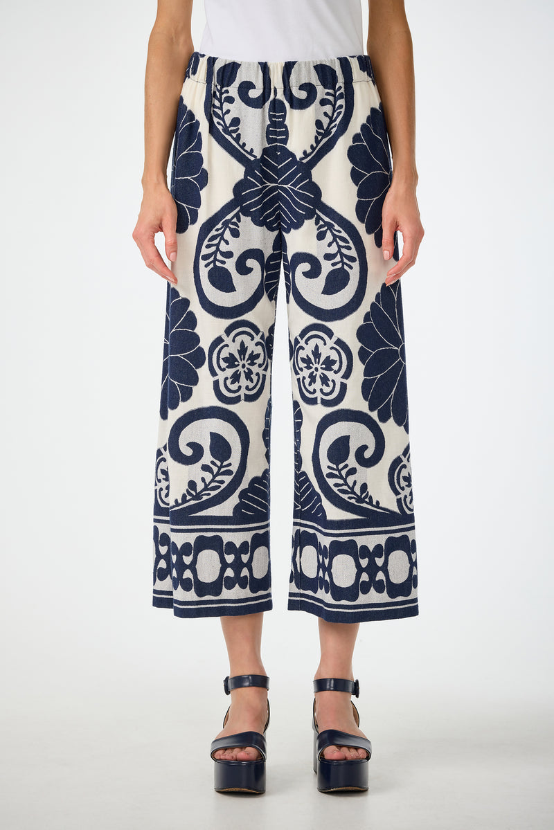 A person wears the Estella 2 Cropped Pants in Fuyuko Fil Coupe—wide-leg, ankle-length pants with bold navy blue floral and geometric patterns—paired with a white top and black platform sandals against a plain background.