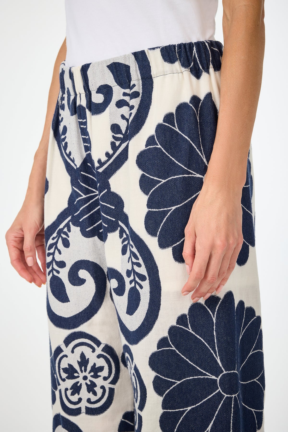 A person wears the Estella 2 Cropped Pants - Fuyuko Fil Coupe, featuring a blue floral and swirl pattern on white with an elastic waistband, paired with a white top and standing with one arm at their side.