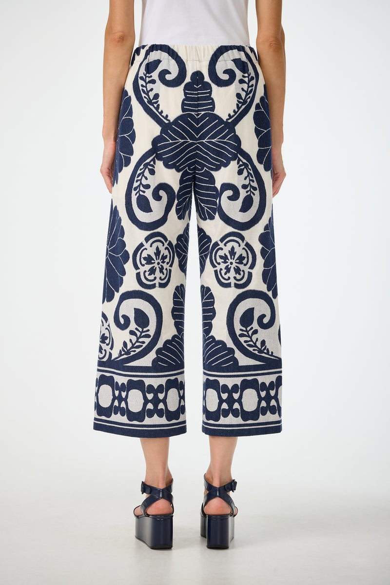 A person is seen from the back wearing Estella 2 Cropped Pants - Fuyuko Fil Coupe, featuring wide-leg, cropped white pants with bold navy blue floral and geometric patterns, paired with black platform sandals.