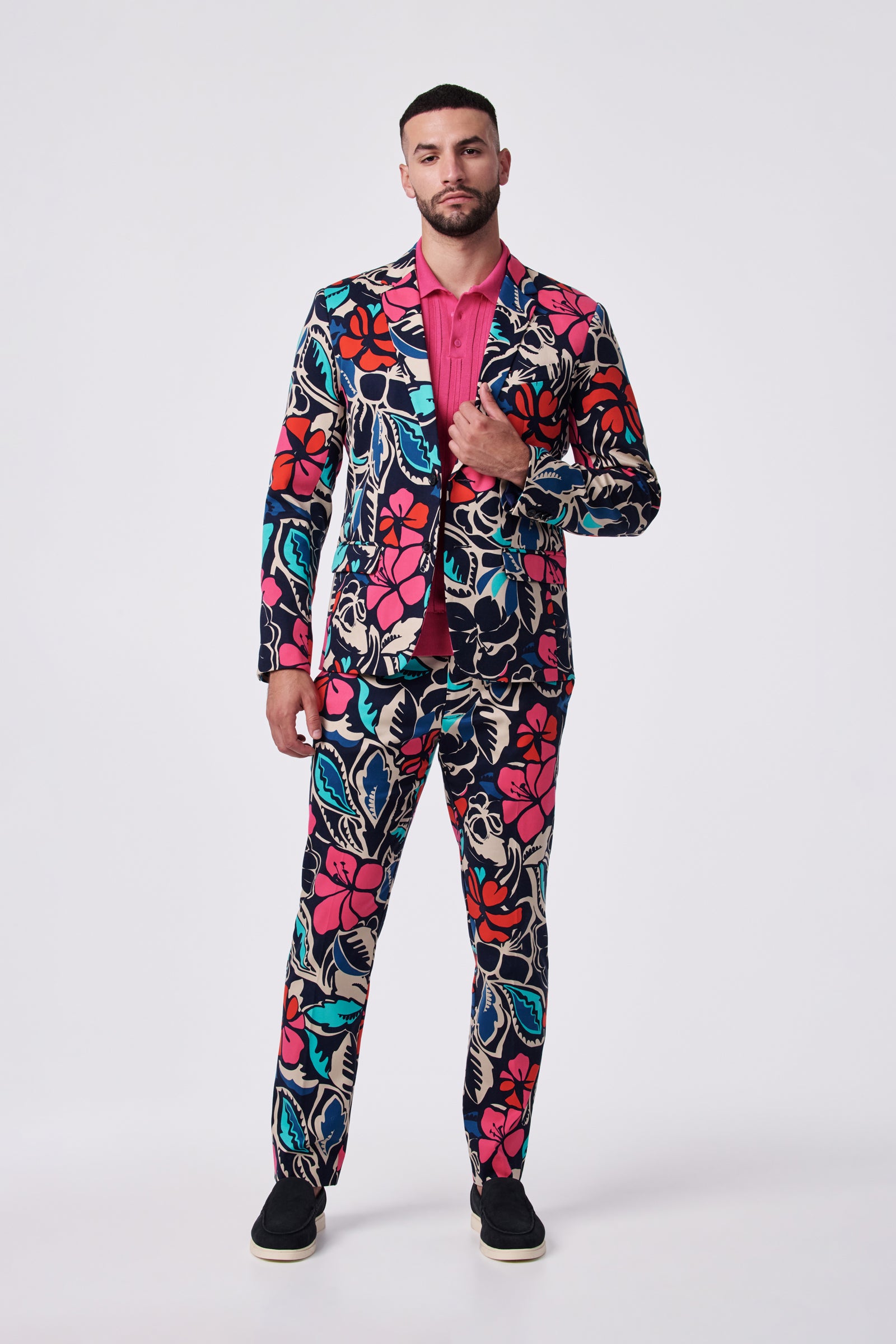 A man with short hair and a trimmed beard stands against a plain background, adjusting his Thurston Blazer—a colorful floral suit jacket in blue, red, pink, and cream—worn over a pink shirt.