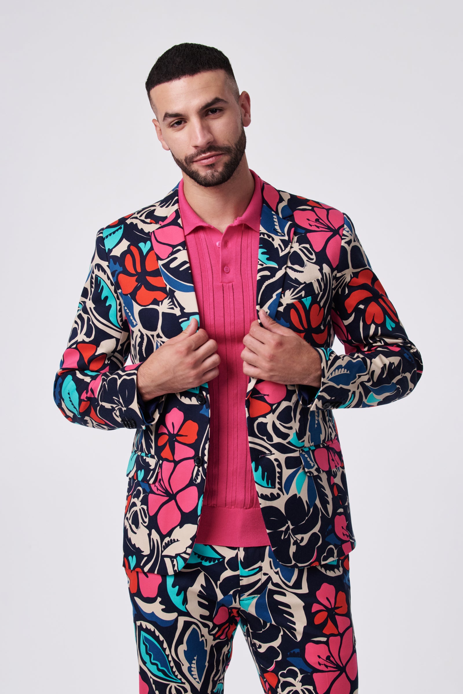 A man poses confidently against a plain background wearing the Thurston Blazer, which features bold, colorful floral patterns, paired with a bright pink collared shirt. He holds the jacket lapels and looks directly at the camera.