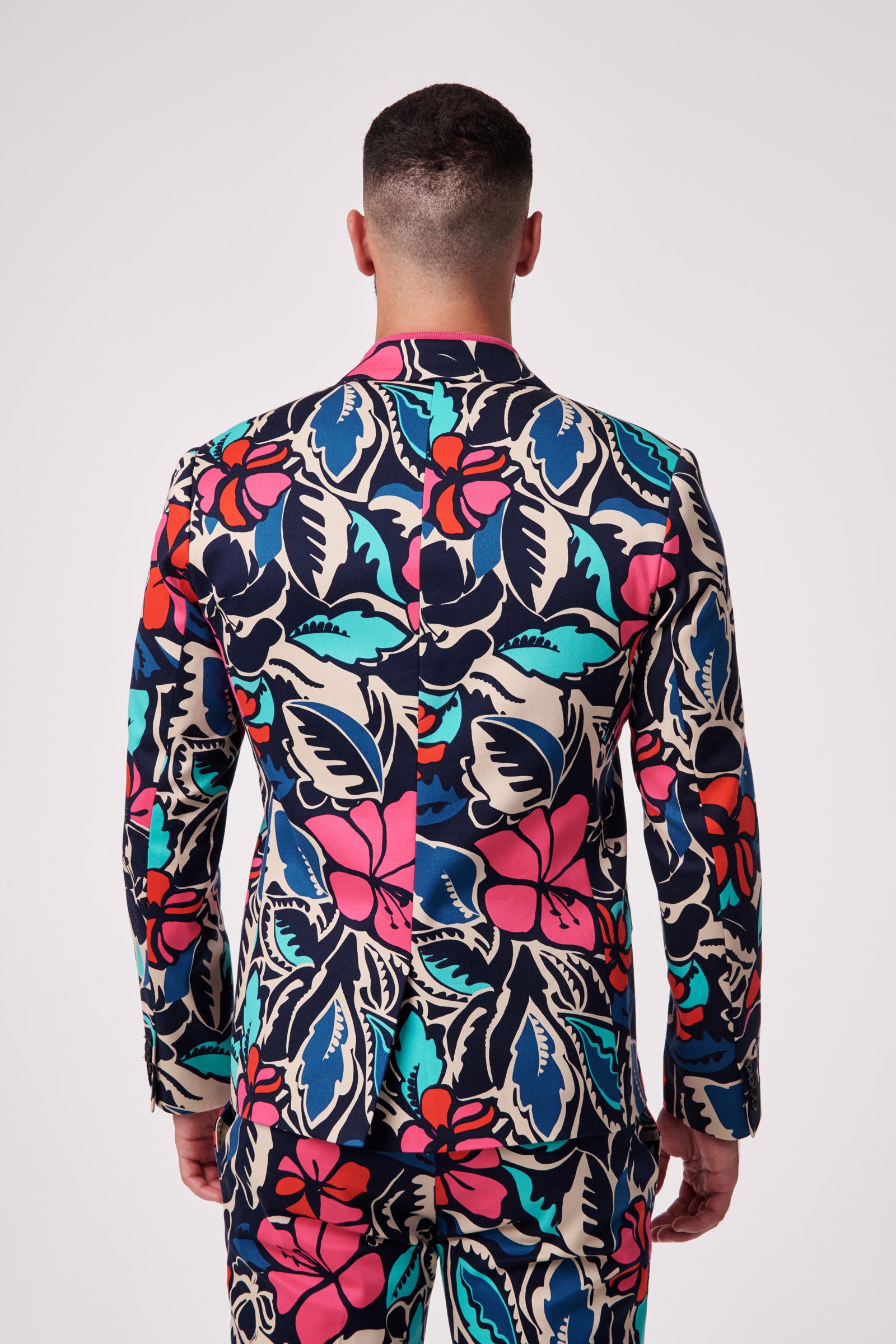 A man faces away, wearing the Thurston Blazer—a suit with bold pink, red, turquoise, and navy floral patterns on beige. The look features a matching jacket and pants against a plain white background.