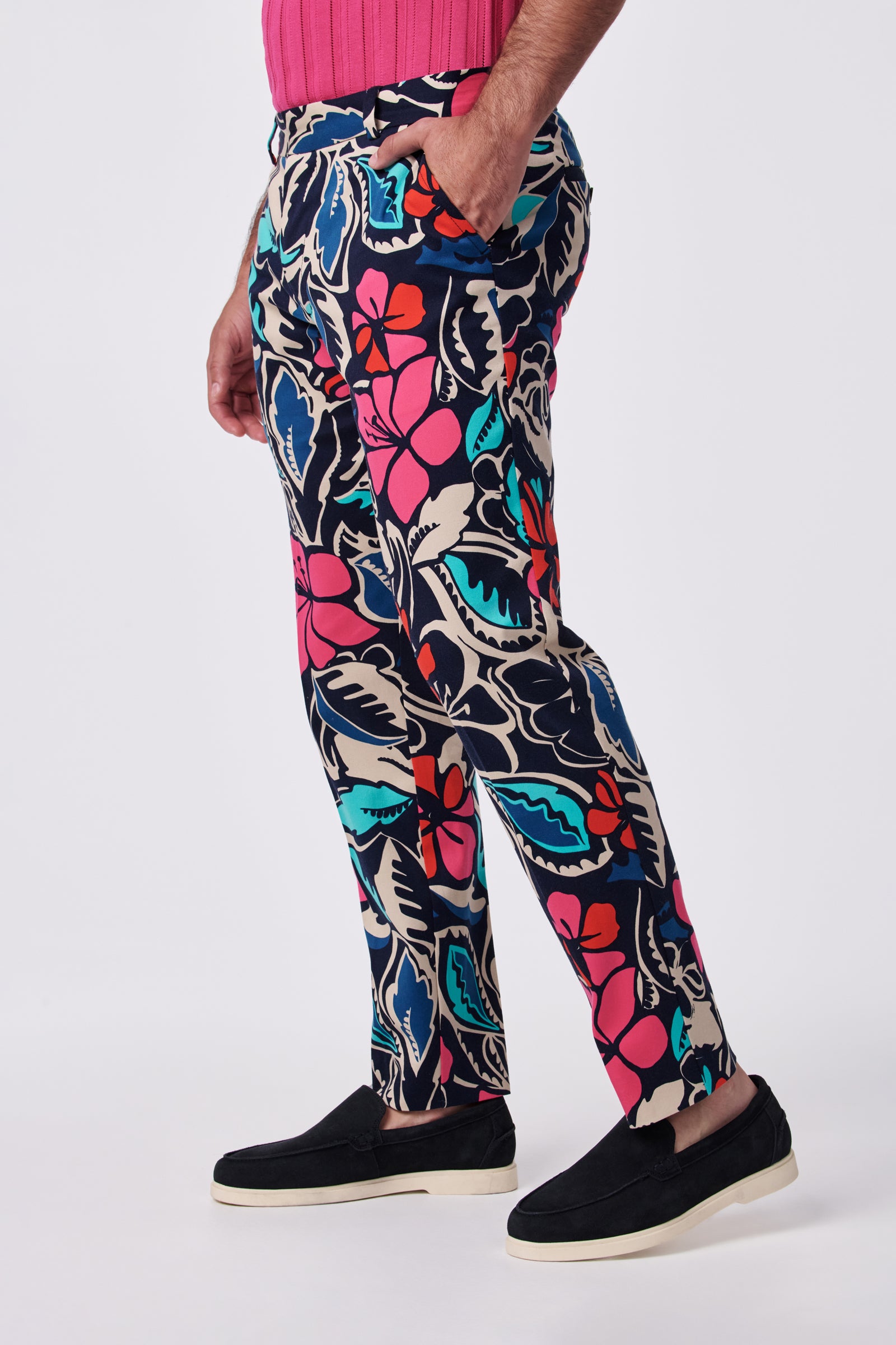 A person stands sideways wearing Clyde Slim Trouser in dark blue with a bold floral pattern in red, pink, teal, and beige, paired with a pink ribbed top and black slip-on shoes with white soles.