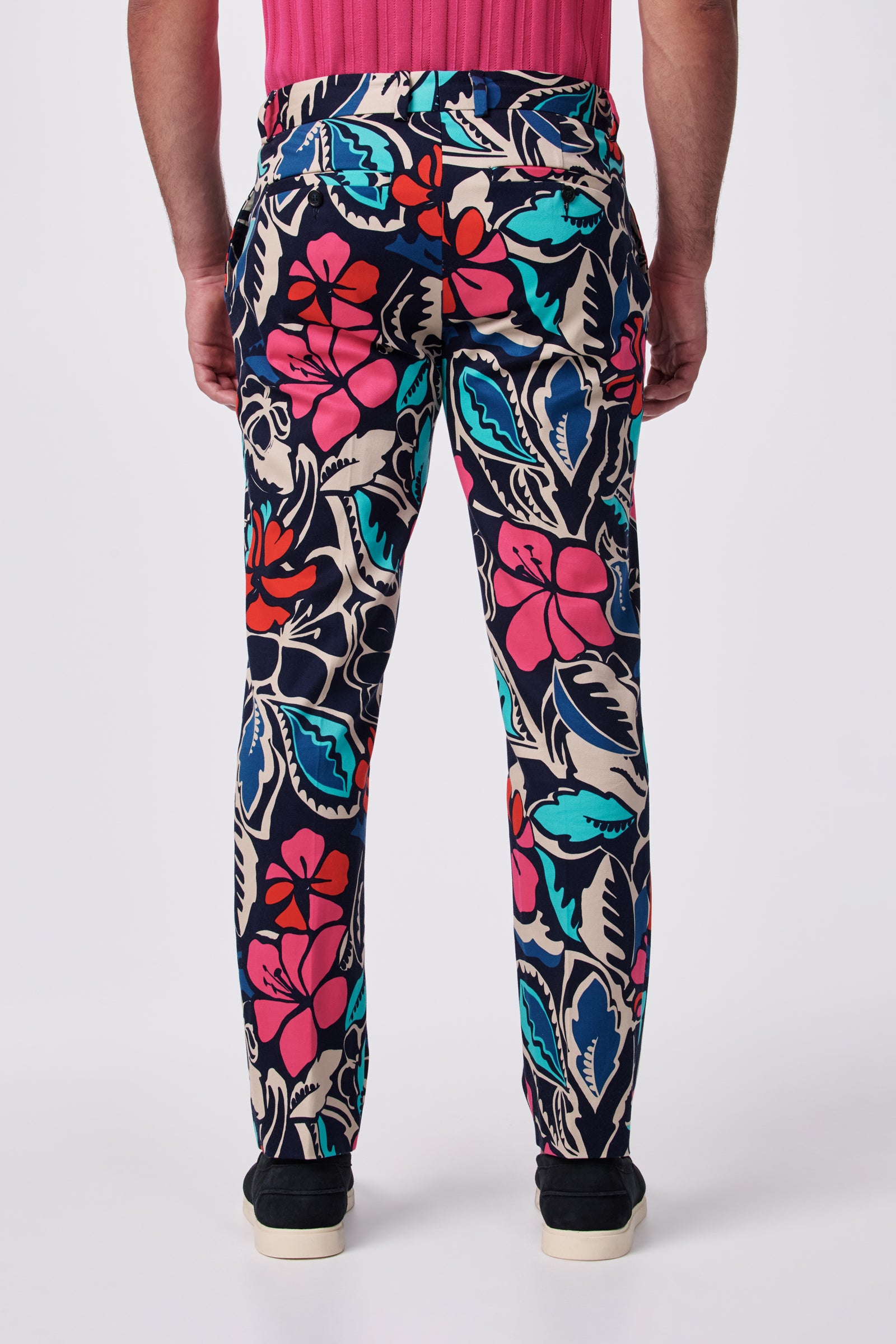 A person stands facing away, wearing the Clyde Slim Trouser featuring colorful red, pink, blue, and teal floral patterns, paired with a pink shirt and black shoes against a plain white background.