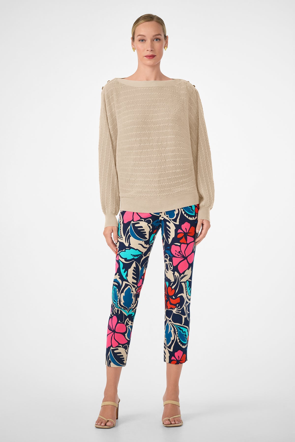 A woman with light skin and blonde hair pulled back faces forward, wearing the beige textured Ima Sweater, bold floral pants in blue, red, pink, and black, and beige high-heeled sandals.