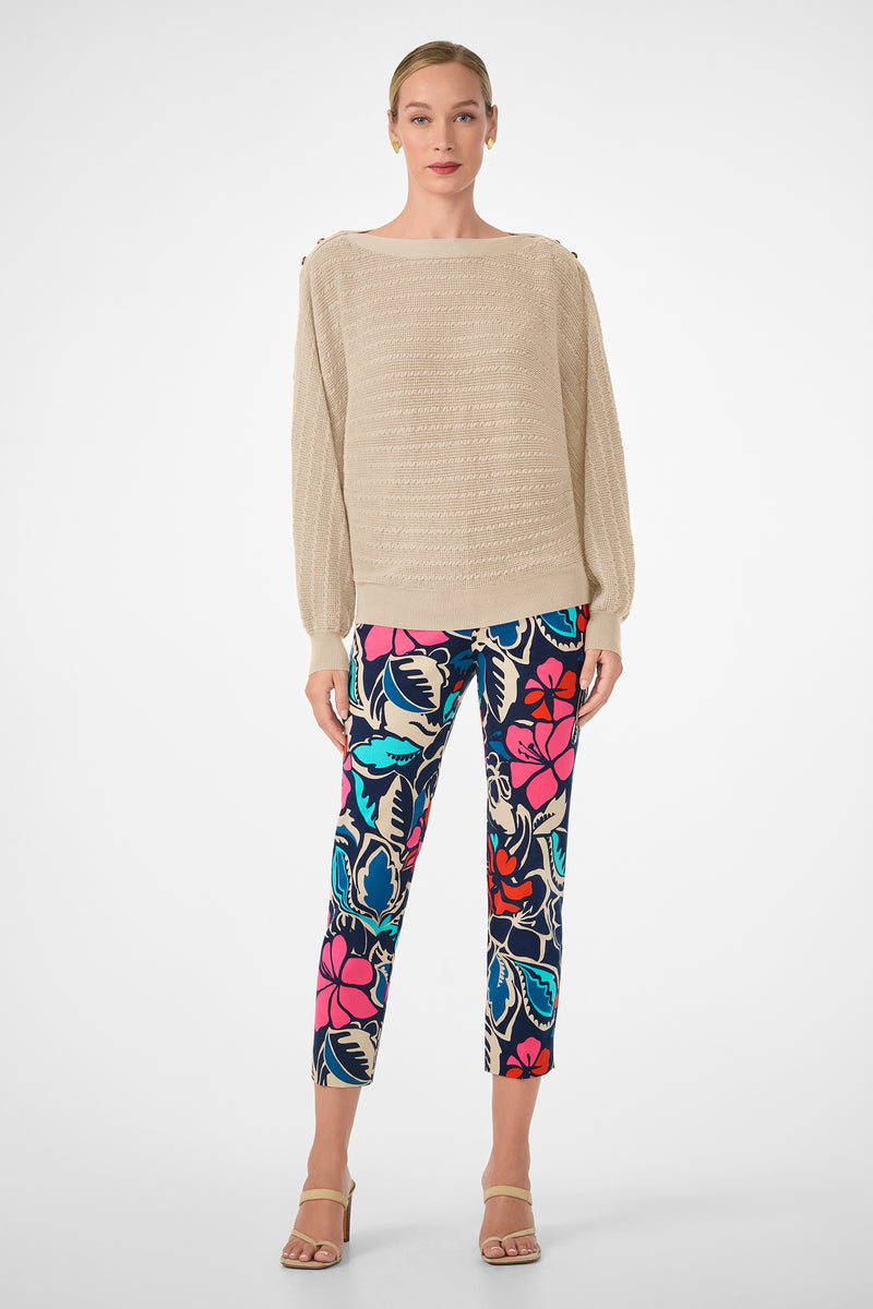 A woman with light skin and blonde hair pulled back faces forward, wearing the beige textured Ima Sweater, bold floral pants in blue, red, pink, and black, and beige high-heeled sandals.