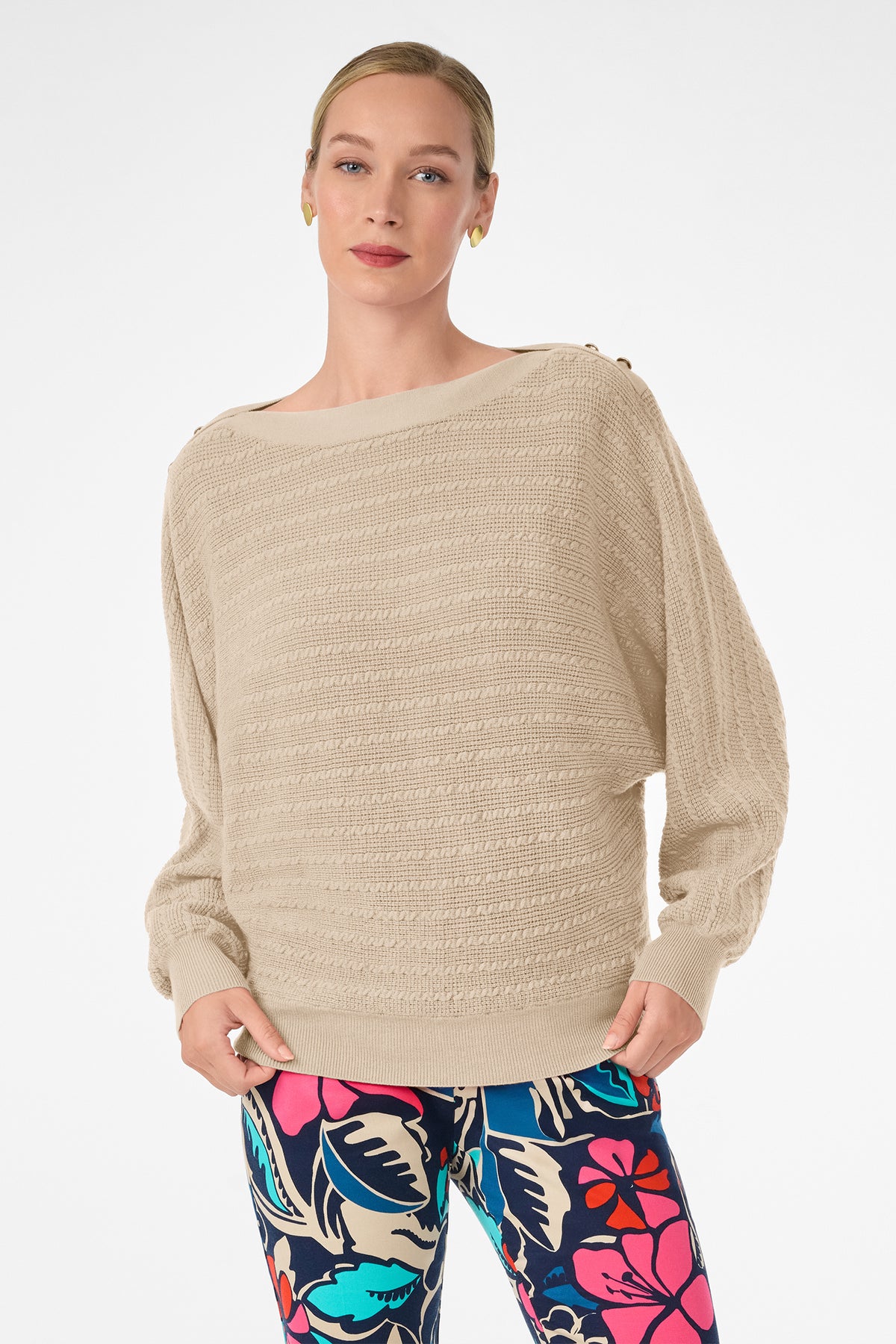 A woman with light hair pulled back and a neutral expression stands against a white background wearing the Ima Sweater, a light beige textured long-sleeve design, paired with colorful floral-patterned leggings.