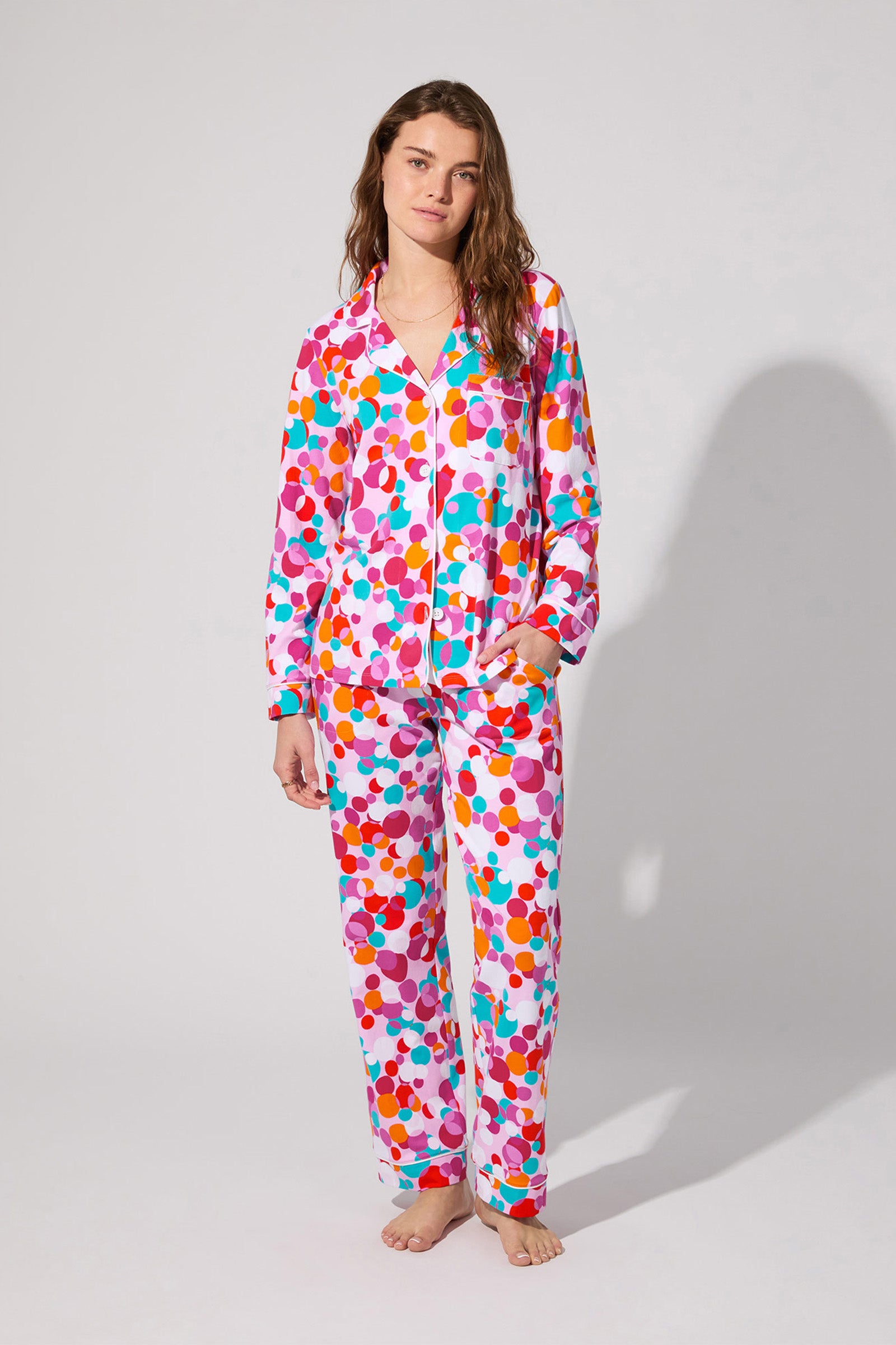 A woman stands barefoot in the Bubble Dots Classic Pj Set—long-sleeved, colorful polka dot pajamas with pants—in shades of pink, red, orange, cyan, and purple against a plain light gray background.