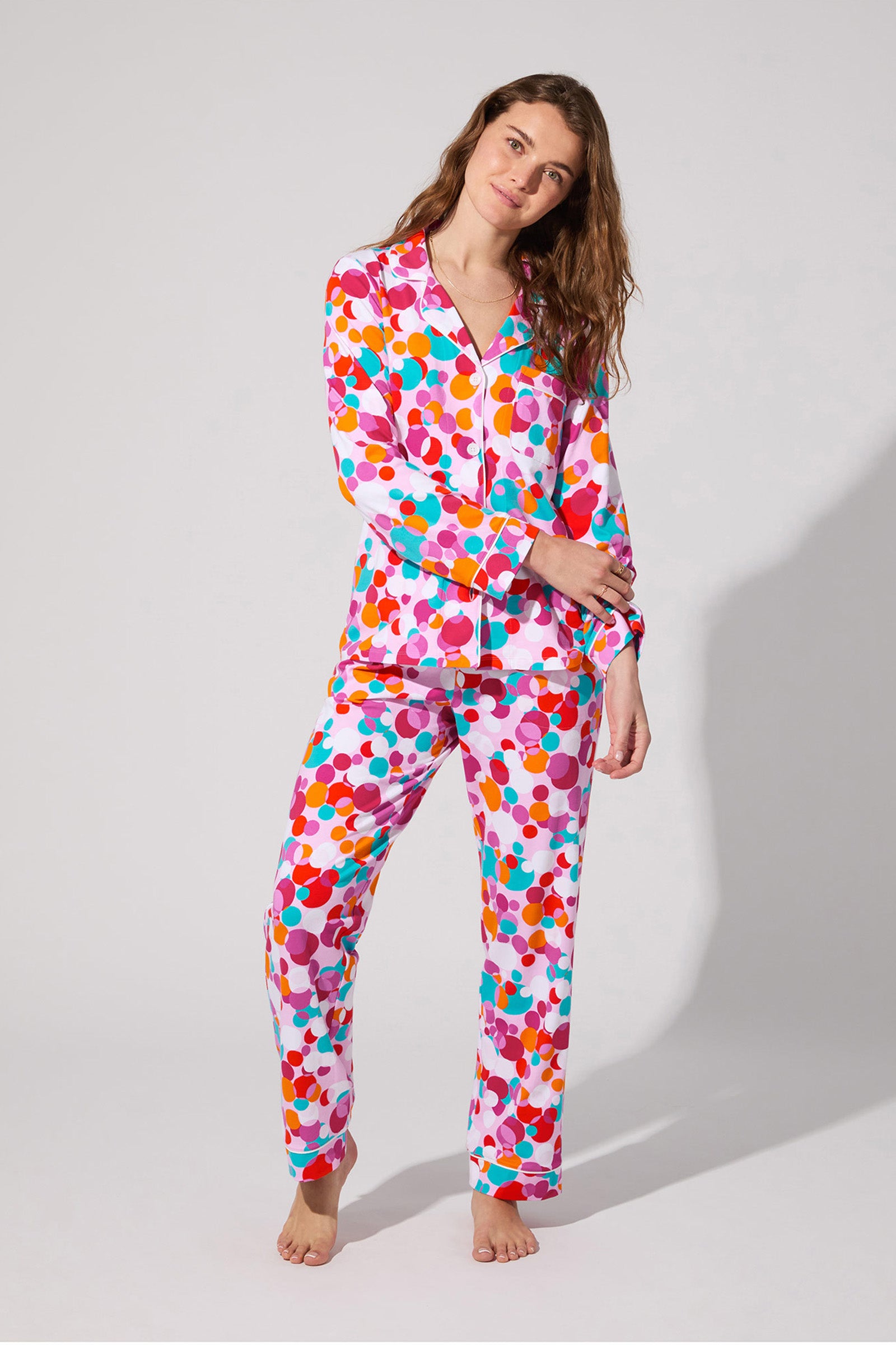 A woman, barefoot and relaxed, wears the Bubble Dots Classic Pj Set featuring a vibrant polka dot pattern in red, pink, purple, blue, and orange as she looks at the camera.
