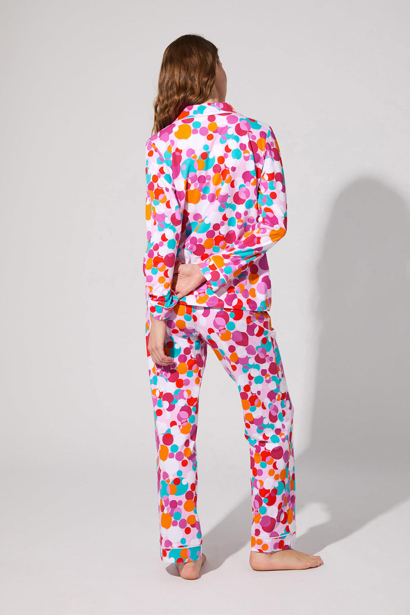 A person with long hair stands barefoot facing away, modeling the Bubble Dots Classic Pj Set—colorful polka-dotted pajamas featuring pink, orange, purple, and blue dots on white—against a plain light gray background.