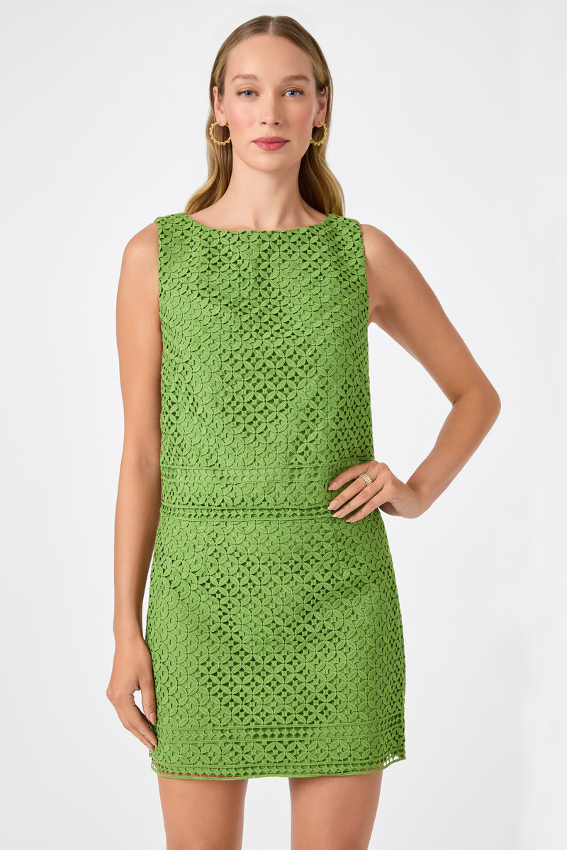 A woman poses against a white background wearing the Cayley 2 Top - Lumi Lace in Sogen Green. The sleeveless top features a crochet pattern and boat neckline. She smiles softly, with one hand on her hip and long, straight hair.