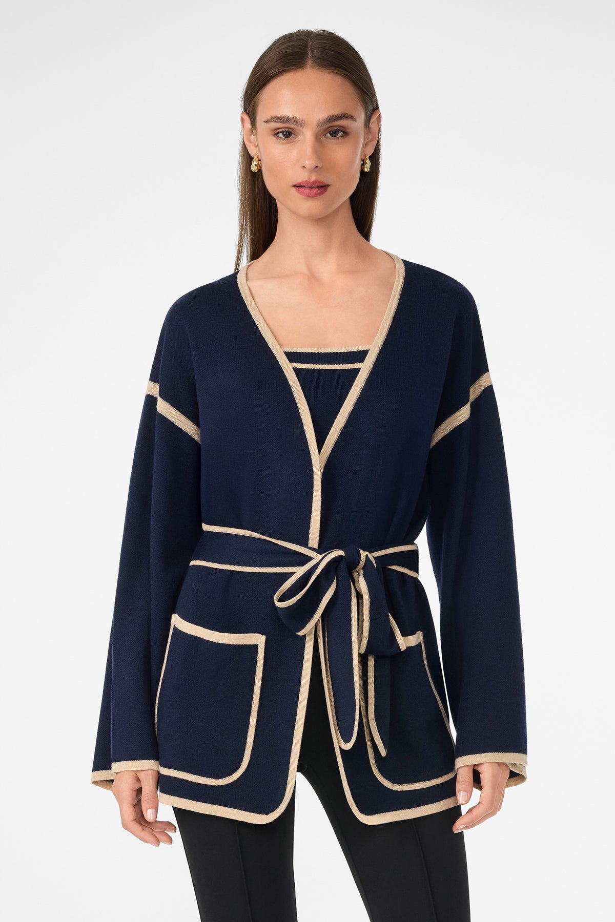 A woman with long straight hair stands against a white background wearing the Wright Wrap Cardigan in navy with beige trim, featuring large front pockets and a tied waist belt, paired with a matching top and black pants.
