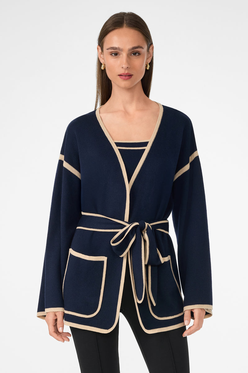 A woman with long straight hair stands against a white background wearing the Wright Wrap Cardigan in navy with beige trim, featuring large front pockets and a tied waist belt, paired with a matching top and black pants.