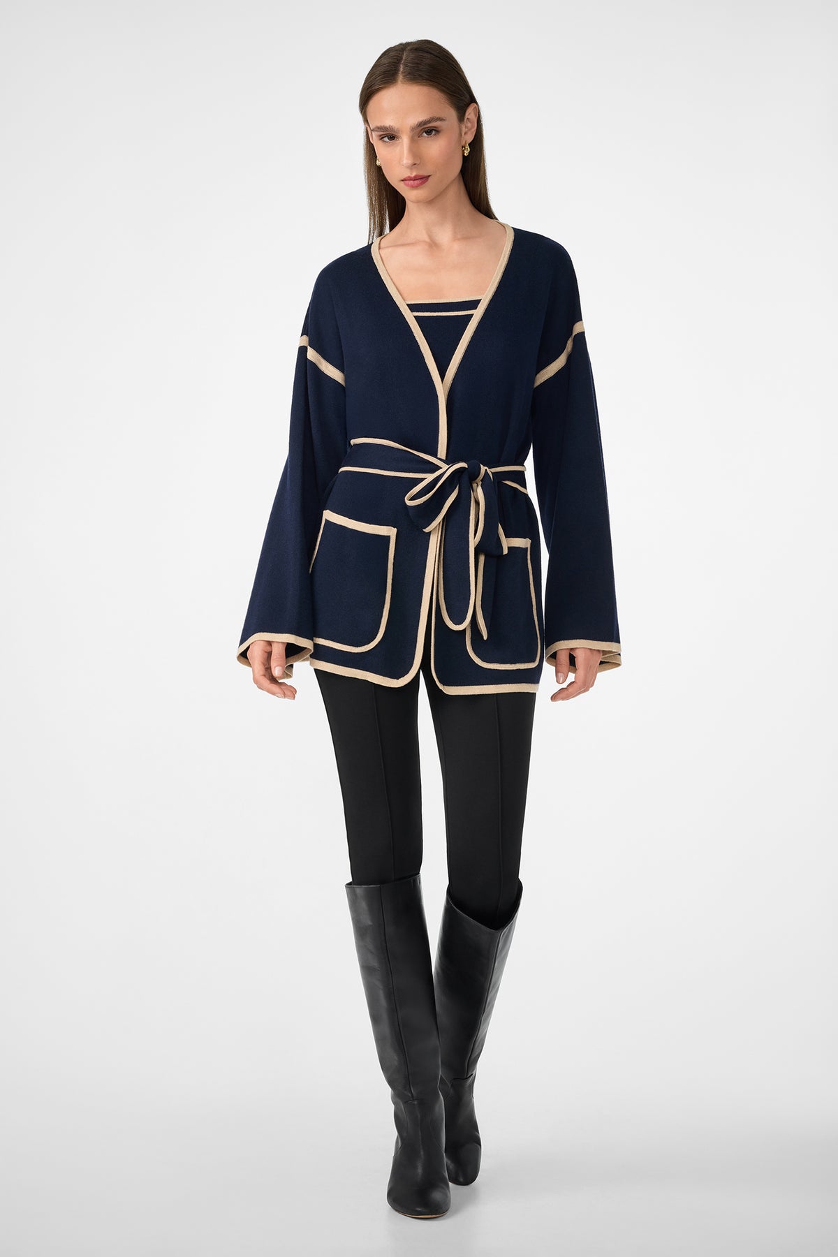 A woman wearing the Wright Wrap Cardigan—navy blue with beige trim and a belt—along with a matching top, black pants, and knee-high black boots, stands against a plain white background.