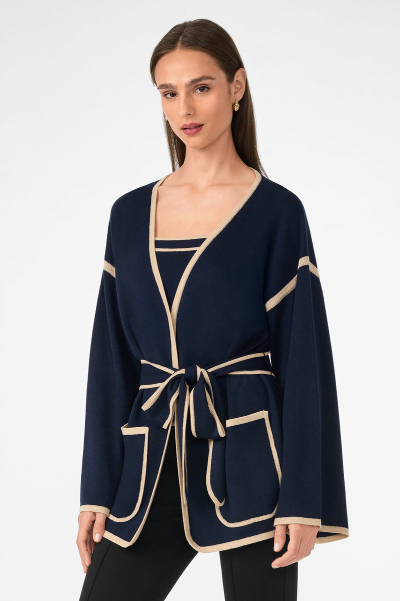 Woman wearing the Wright Wrap Cardigan in navy with beige trim, large front pockets, and a matching waist belt. She has straight brown hair, gold hoop earrings, and stands against a white background.