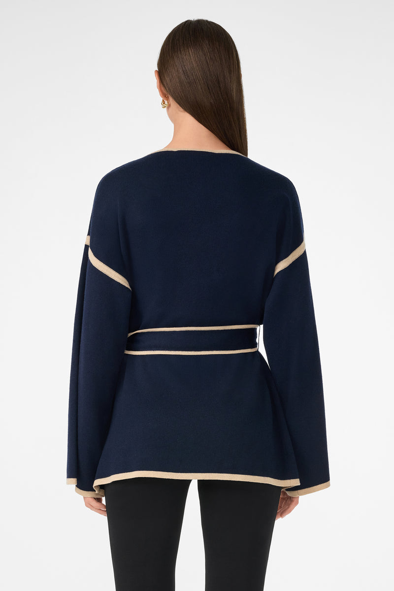 A woman with long brown hair faces away, wearing the Wright Wrap Cardigan in navy blue with beige trim on the sleeves, waist, and hem, paired with black pants against a plain white background.