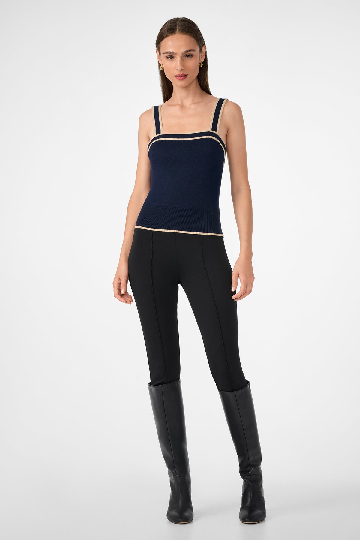 A woman stands against a plain background wearing the Casady Knit Cami, a navy blue square-neck top with beige trim, paired with black fitted pants and knee-high black boots. She has straight brown hair and minimal accessories.