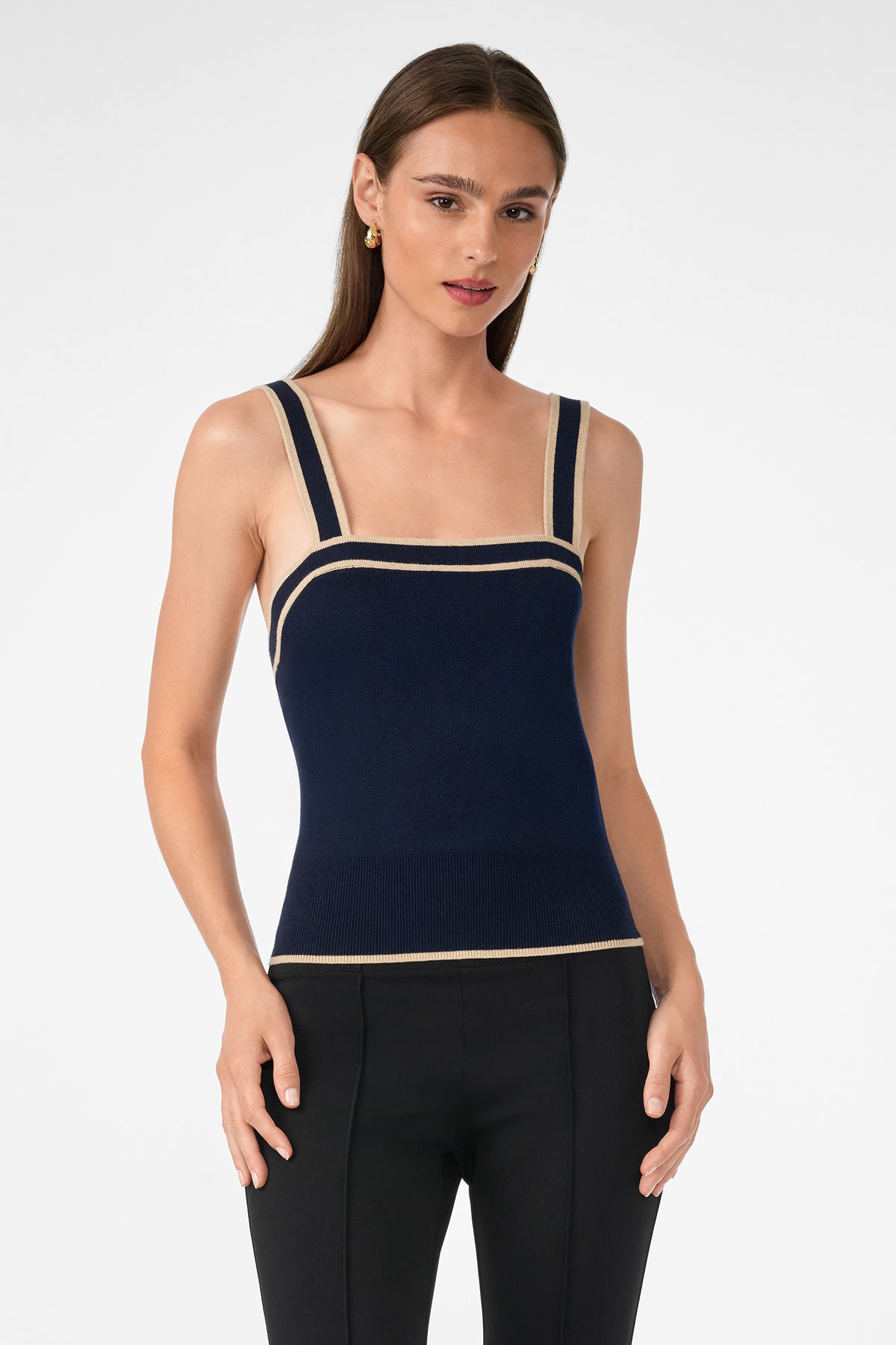 A woman with long brown hair wears the Casady Knit Cami, a navy blue tank top with beige trim, and black pants, standing against a plain white background.