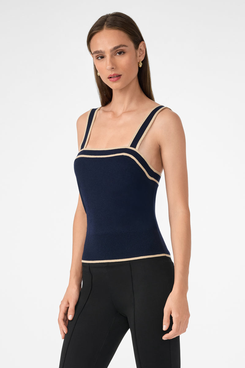 A woman models the Casady Knit Cami—a navy blue sleeveless knit top with beige trim and double shoulder straps—paired with black fitted pants, standing against a plain white background.