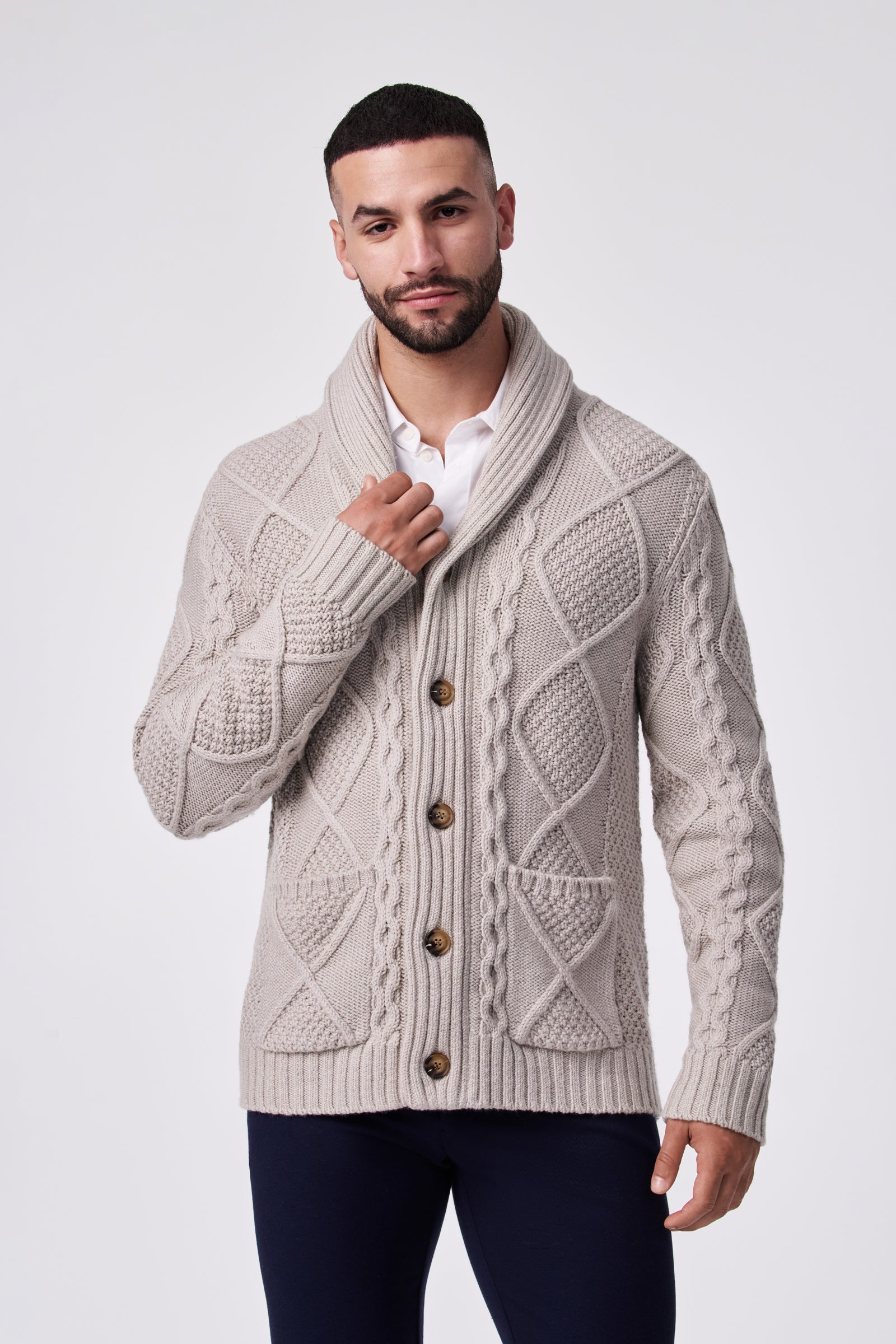 A man with short dark hair and a trimmed beard models the beige Brighton Cardigan, a textured knit style with large buttons, layered over a white collared shirt. He stands against a plain light background, touching the cardigan collar.