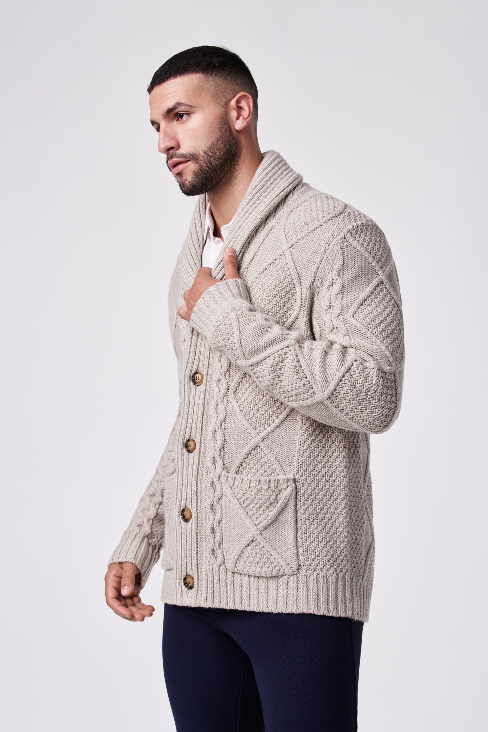 A man with short dark hair and a beard wears the Brighton Cardigan, a beige knit piece featuring large buttons and textured patterns, standing against a plain light background.