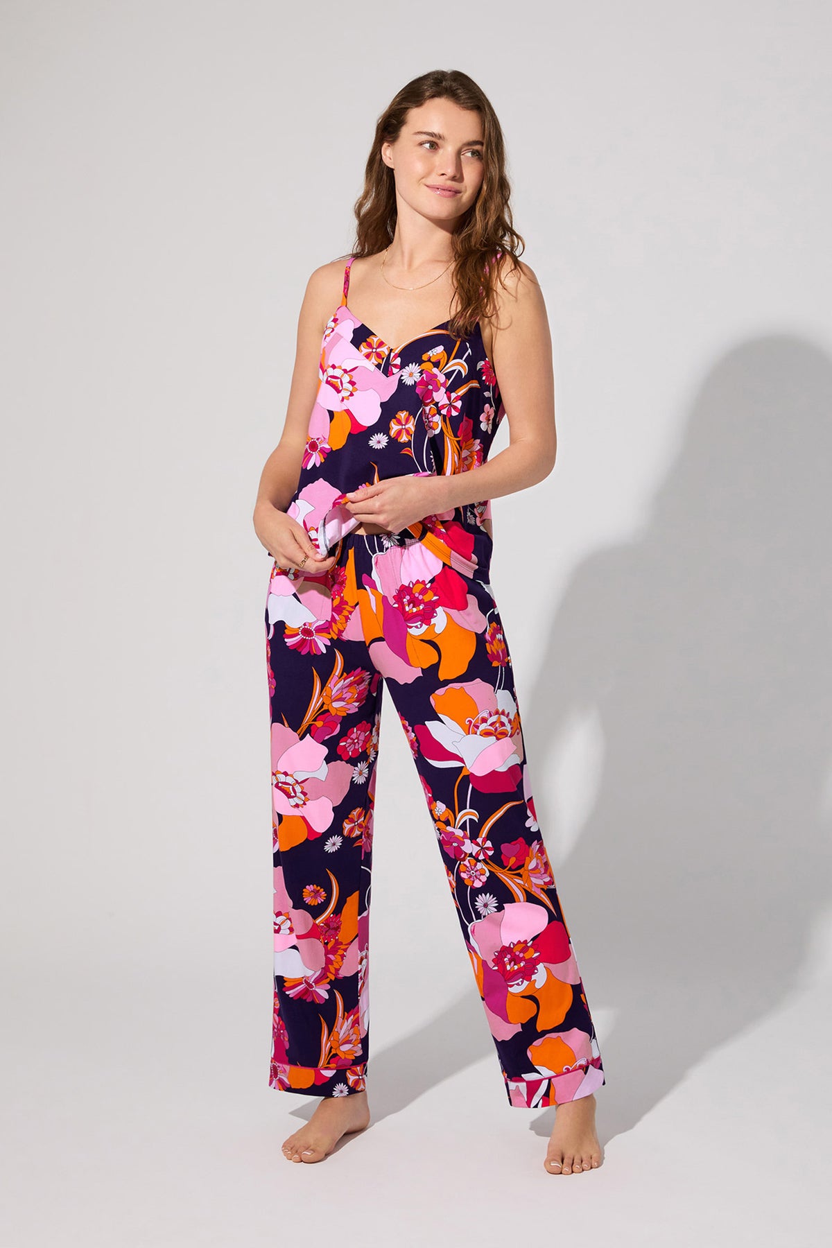 A woman stands barefoot against a plain light background, wearing the Floral Medley Cami Pj Set—a colorful floral pajama set featuring spaghetti straps and wide-leg pants.