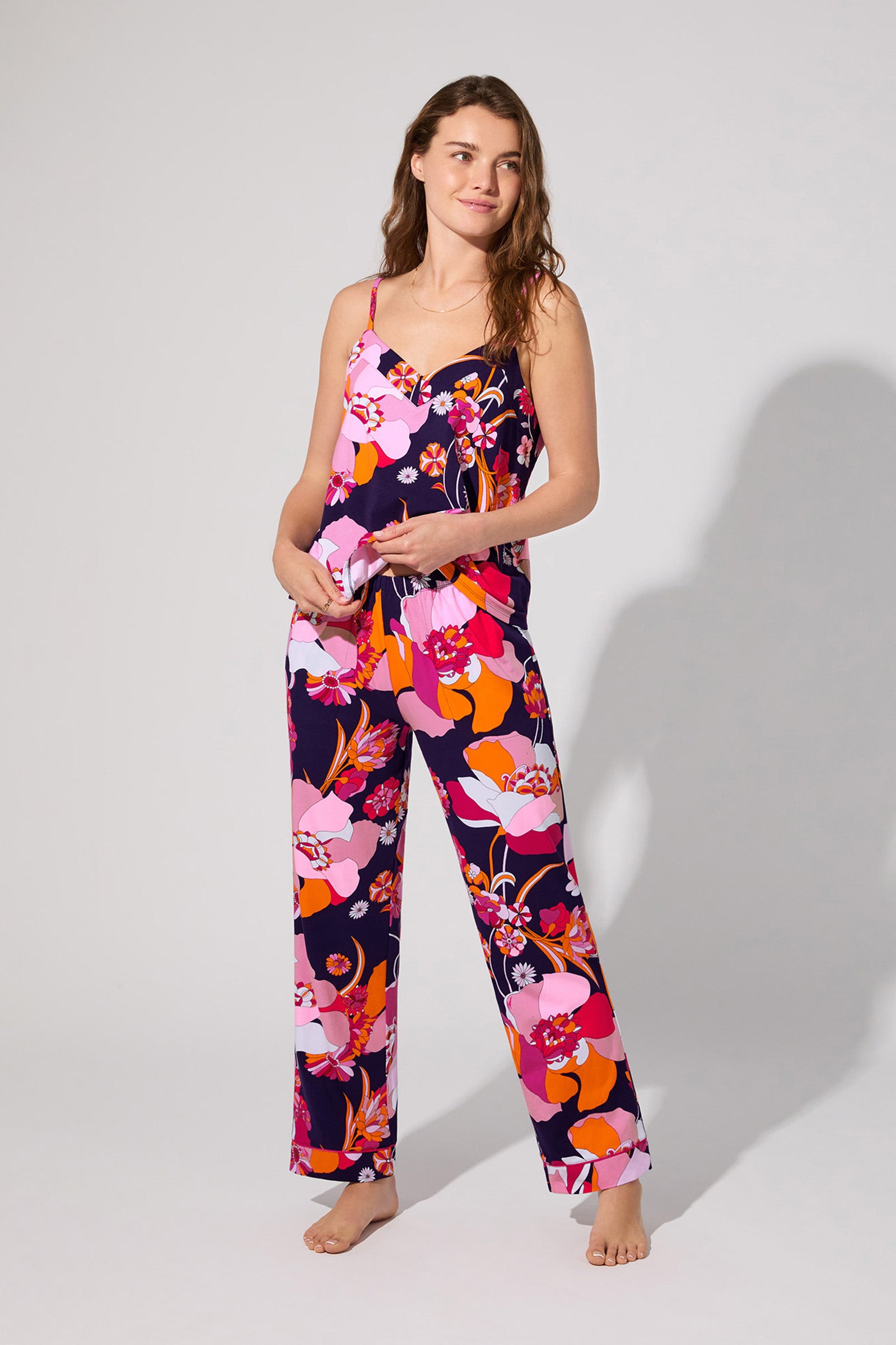 A woman stands barefoot against a plain light background, wearing the Floral Medley Cami Pj Set—a colorful floral pajama set featuring spaghetti straps and wide-leg pants.