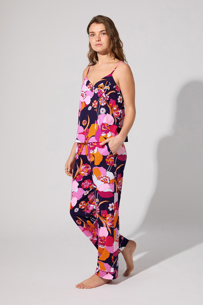 A woman stands barefoot in the Floral Medley Cami Pj Set, featuring a colorful floral print, sleeveless top, and long pants. She poses against a plain background with a relaxed expression, looking slightly to the side.