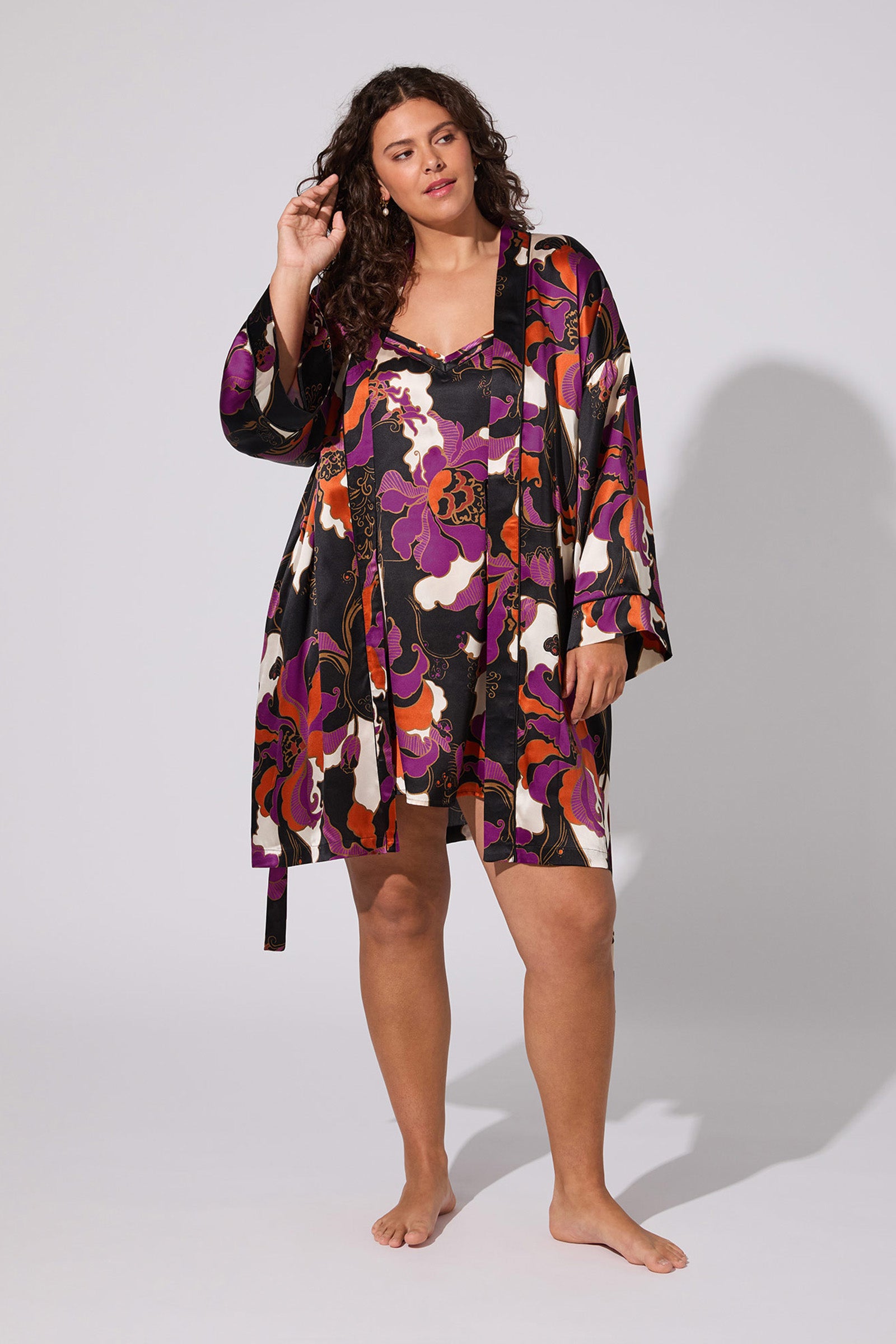 A woman with curly hair models the Fall Floral Silk Robe, a colorful, floral-patterned satin piece in purple, red, orange, and black hues. She stands barefoot against a simple light background.