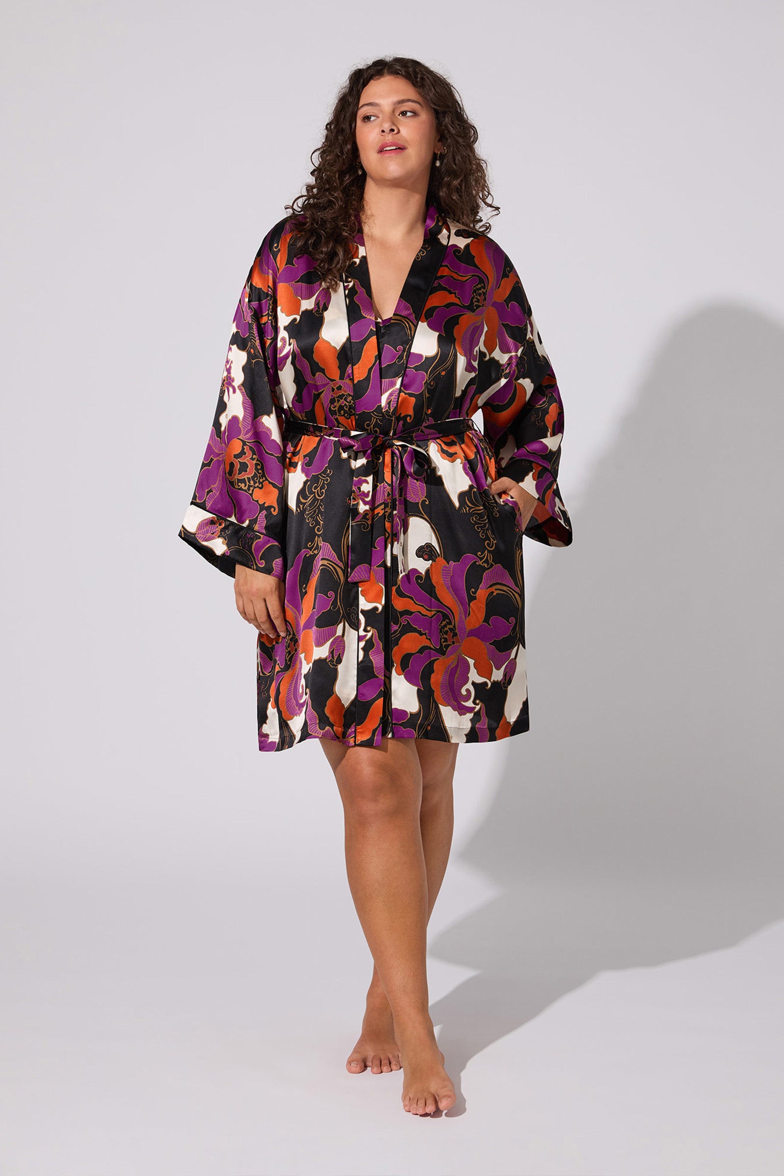 A woman with curly hair models the Fall Floral Silk Robe—a knee-length, long-sleeve piece with a bold abstract floral print in purple, orange, pink, black, and white. She stands barefoot against a plain light gray background.