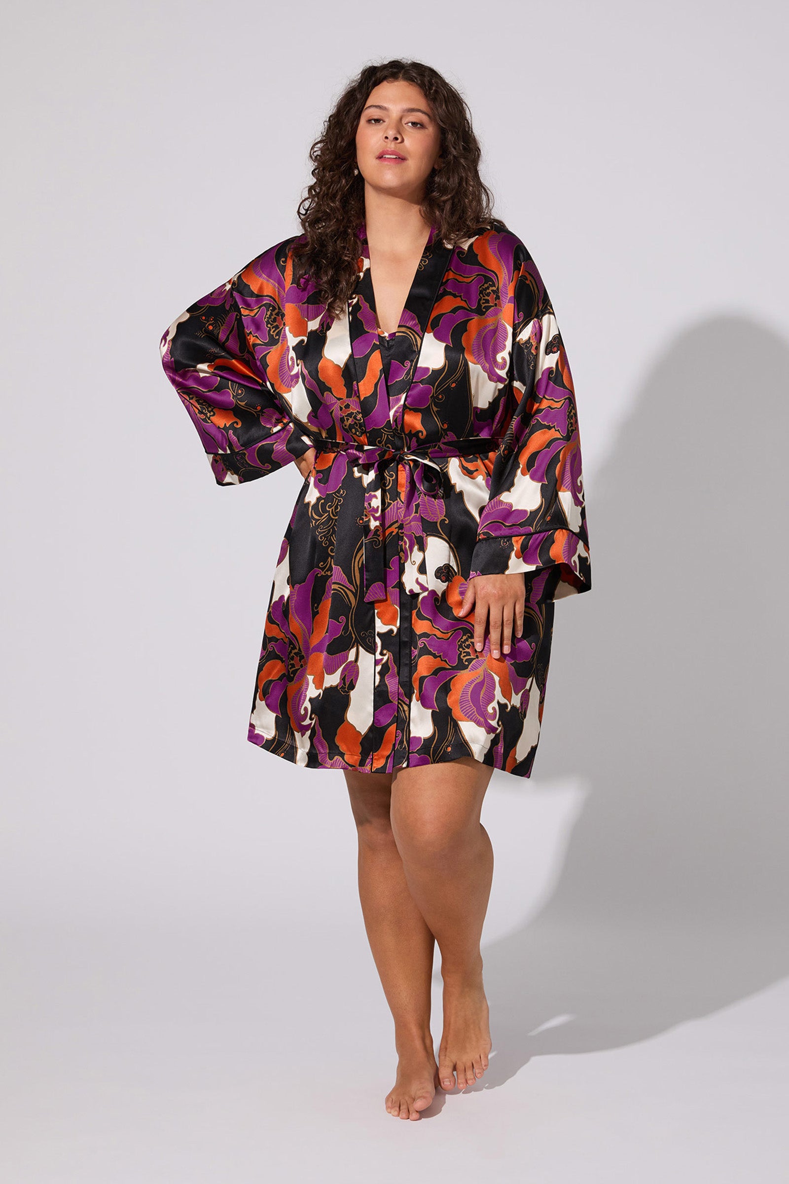 A woman with curly hair stands barefoot against a plain light background, wearing the Fall Floral Silk Robe—an elegant, colorful satin robe with a vibrant abstract pattern and matching belt.