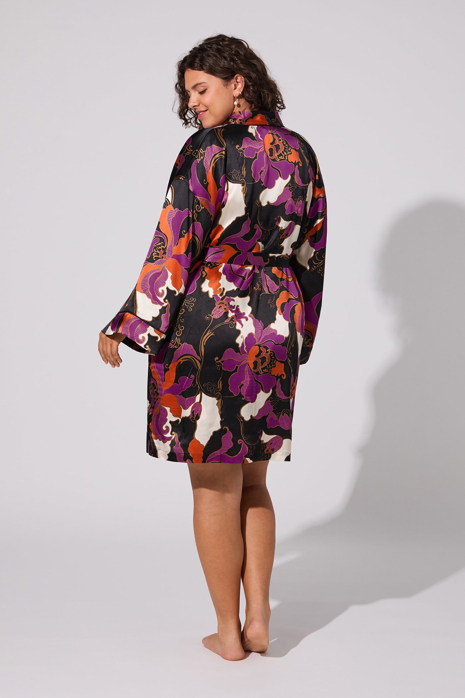A woman with curly hair stands barefoot and smiling, shown from the back against a plain light background, wearing the Fall Floral Silk Robe featuring vibrant purple, orange, and black patterns in a kimono-style design.