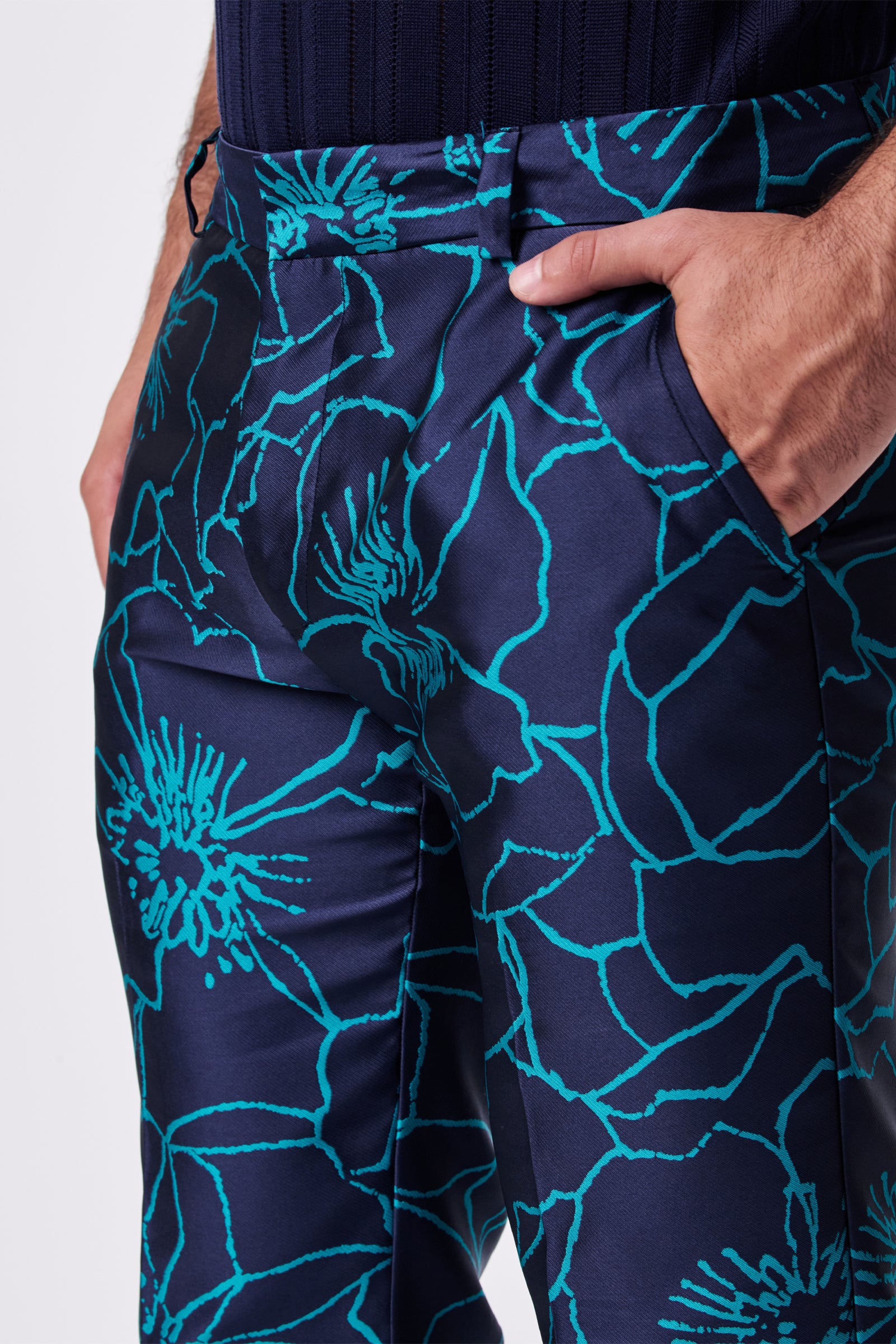 A person wearing the Clyde Slim Trouser in dark blue with a bold turquoise floral pattern stands with one hand in their pocket, highlighting the trouser's striking design and textured fabric.